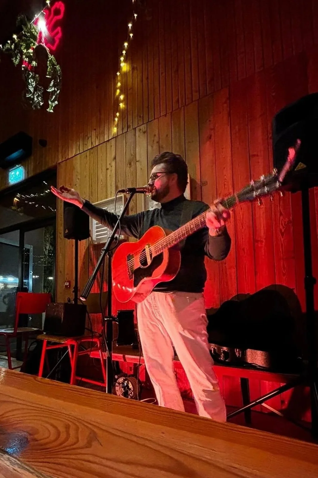 A man performing with a guitar on stage, gesturing with one hand, in a cozy venue with wooden walls and warm lighting, decorated with string lights and holiday decorations.