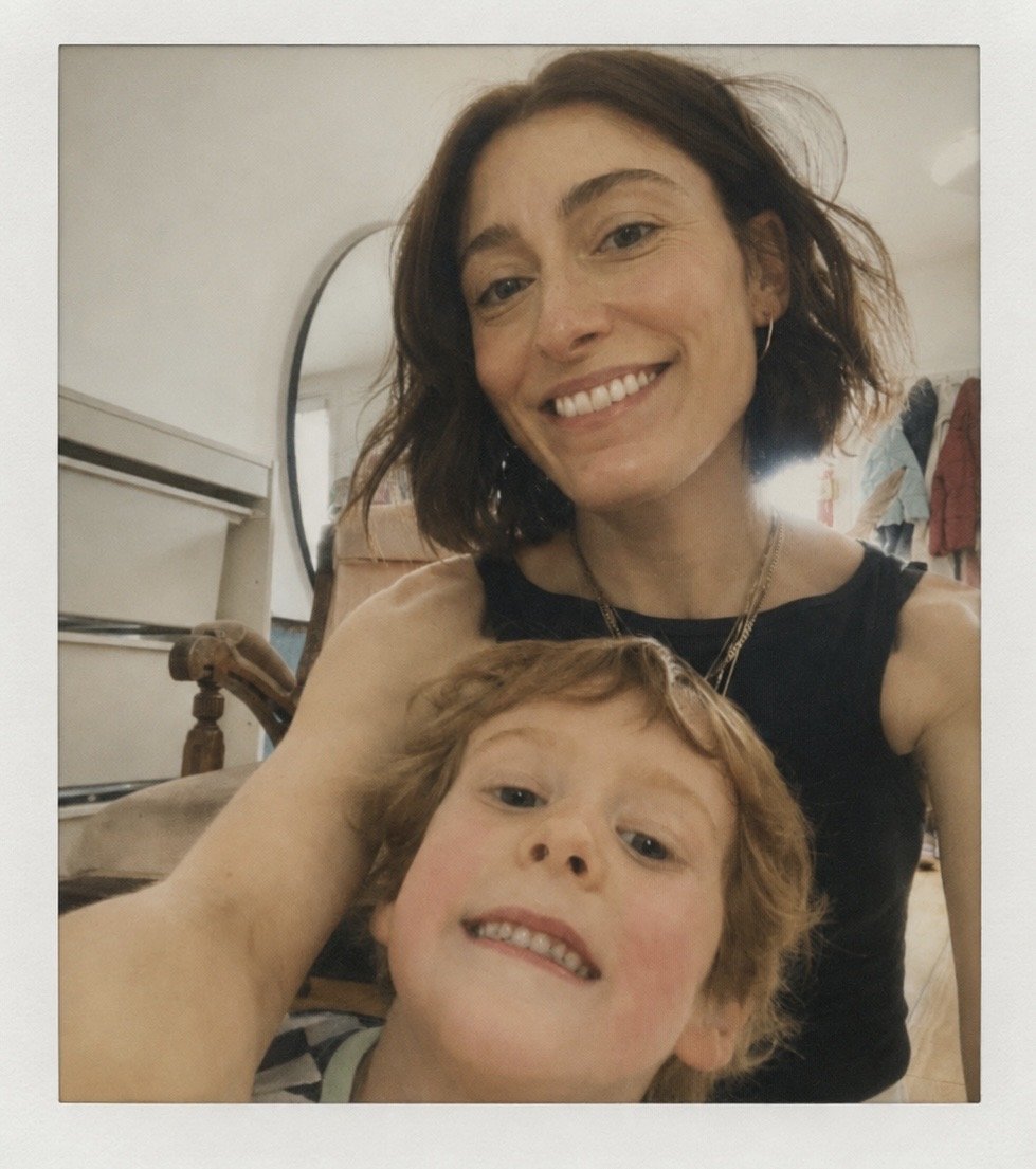 A woman with short brown hair and a boy with curly red hair taking a selfie indoors. The woman is smiling and wearing a black top, while the boy is smiling with his teeth showing. There is a mirror and some clothing in the background.