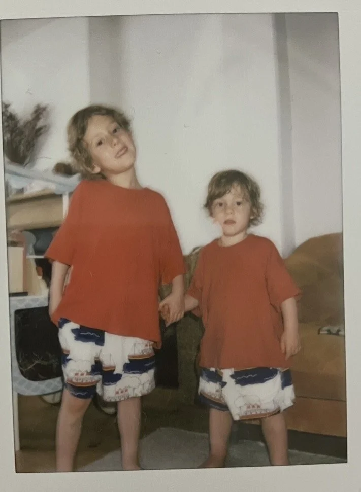 Two young children with curly hair wearing orange T-shirts and beach shorts holding hands in a living room with a bookshelf, plants, and a brown couch.