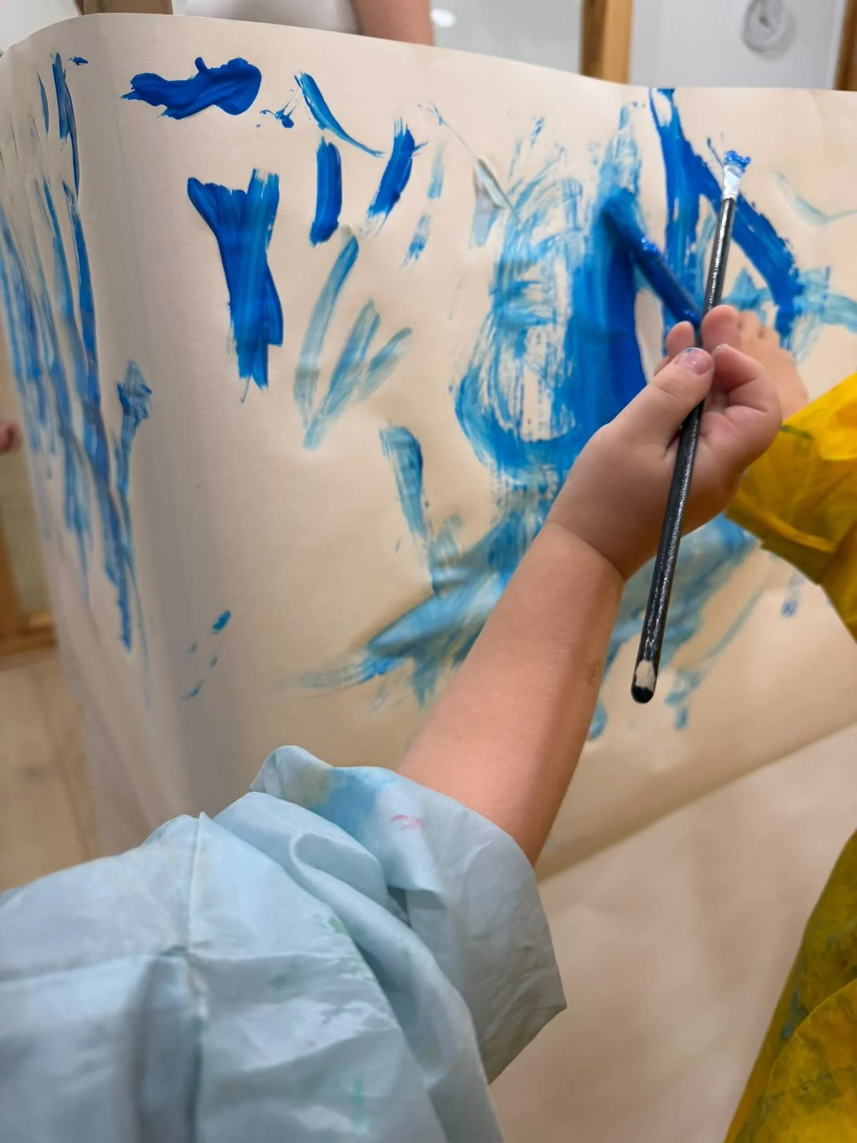 Child painting with blue paint on a large sheet of paper, using a brush, wearing a light blue smock.