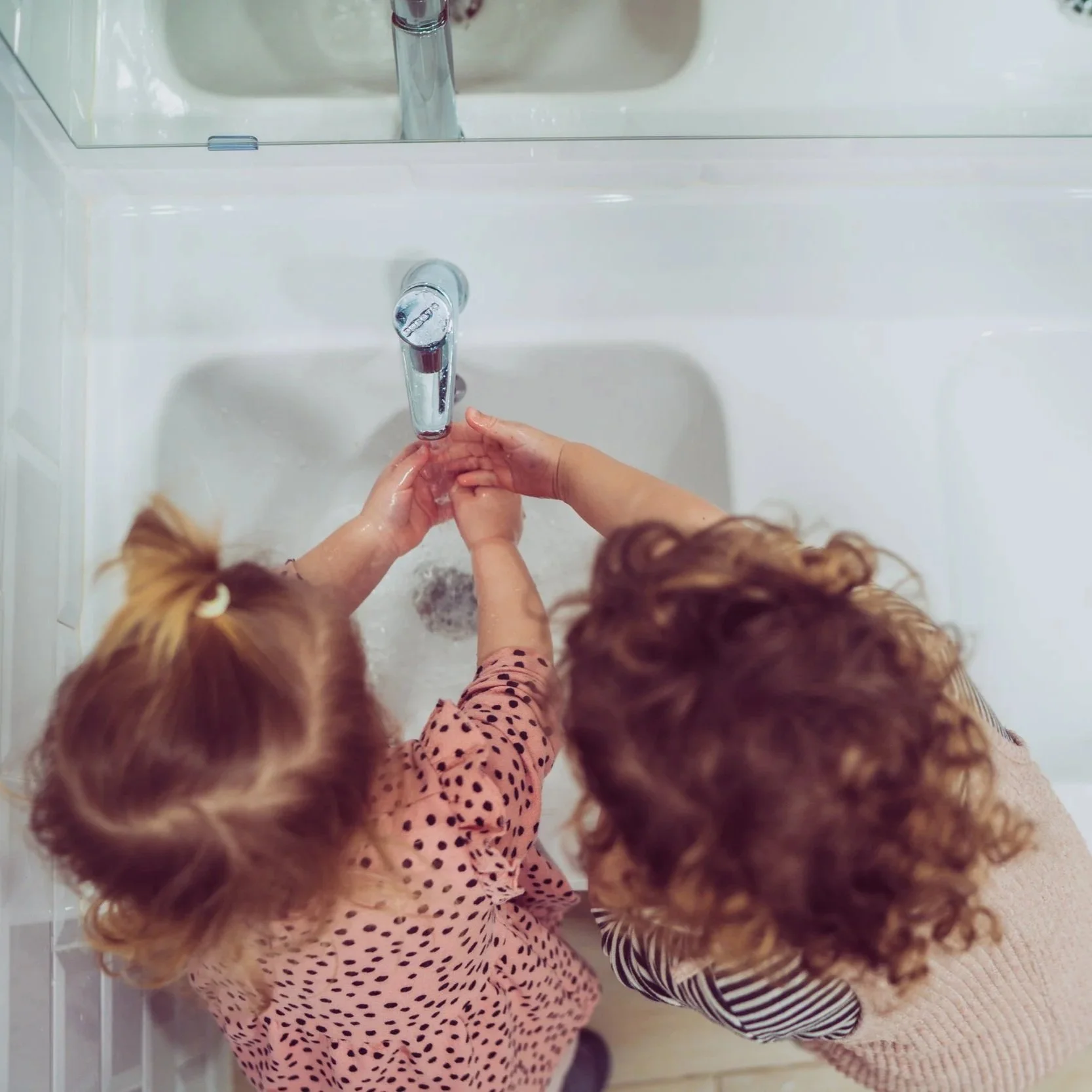 Two children with curly red hair washing hands in a white sink with a mirror above it.
