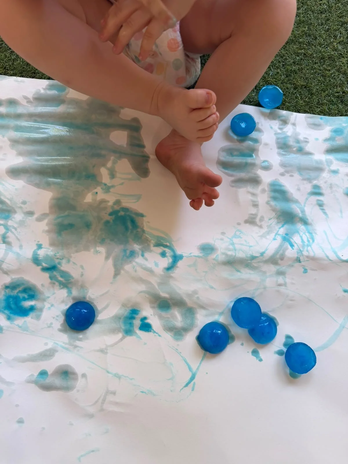 Child engaging in fingerprint and painting activity with blue paint on paper, outdoors on grass.
