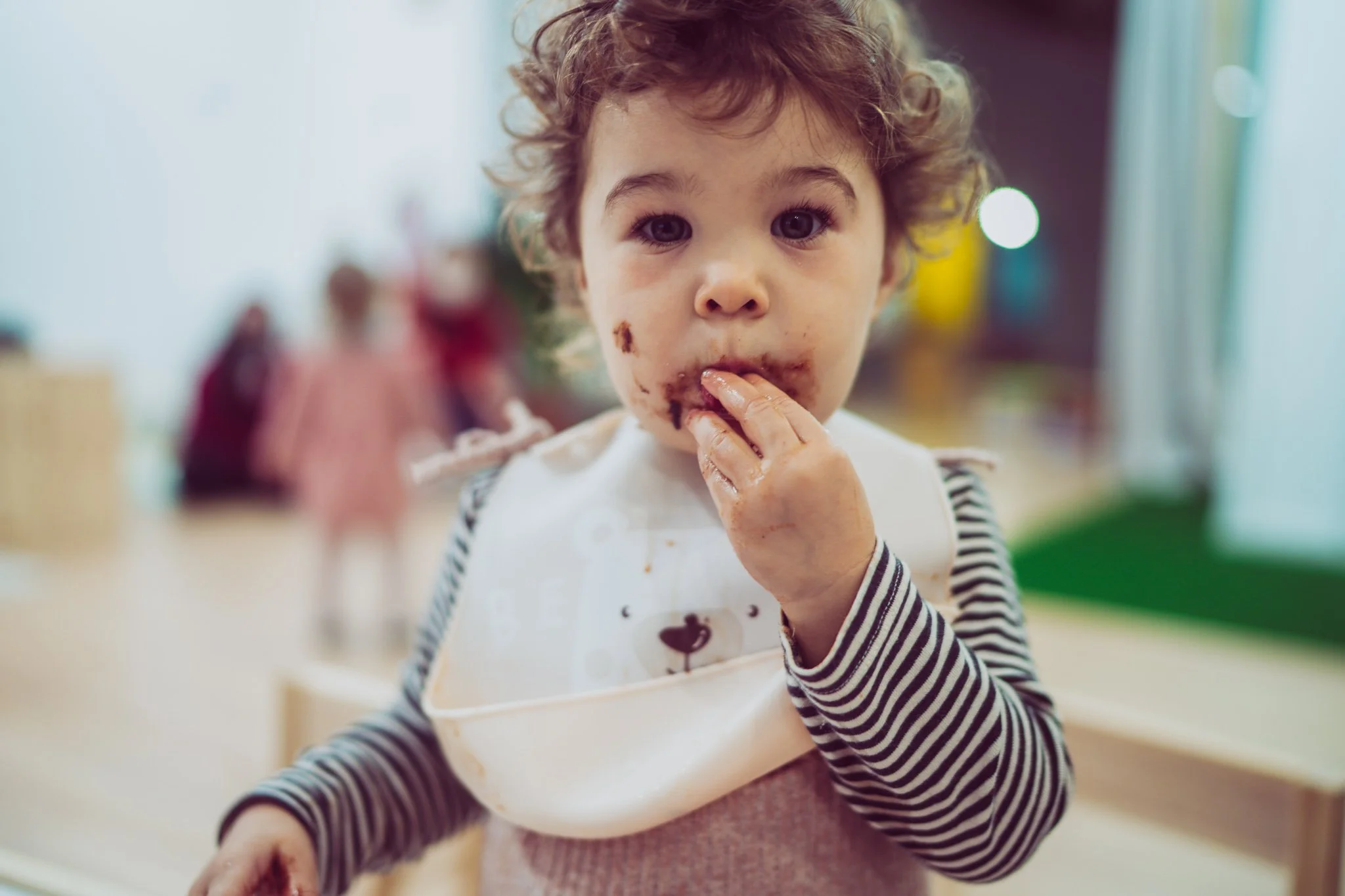 A young child with curly hair and light skin, wearing a striped long sleeve shirt and a white bib with a bear face, is eating a chocolate dessert. The child has some chocolate on their face, fingers, and lips, and appears to be enjoying their messy snack.