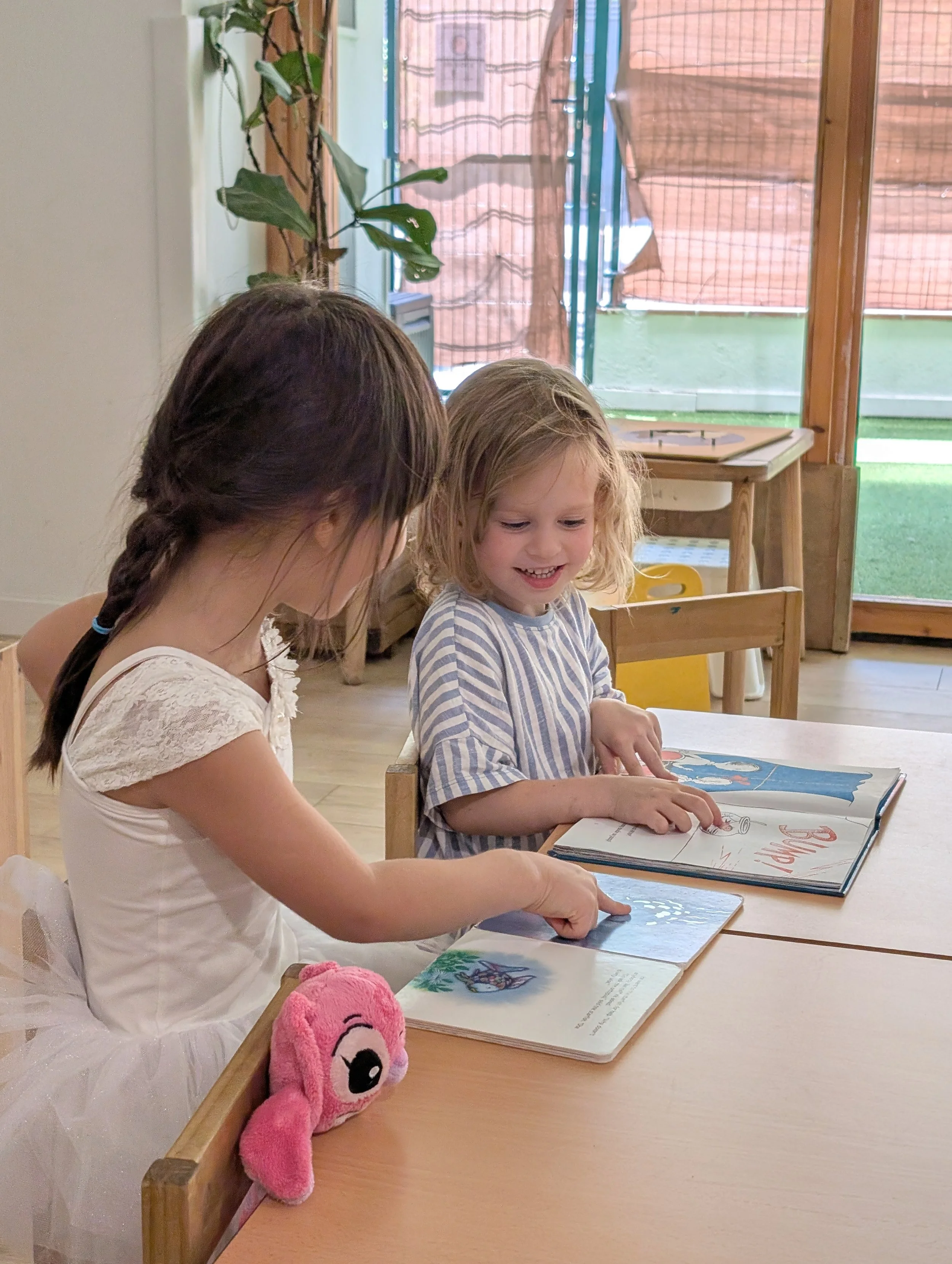 Two young girls sitting at a table, looking at picture books and smiling. One girl has dark hair in a braid, and the other has blonde, curly hair. There is a pink plush toy on the table with a unicorn face. In the background, there is a window with s