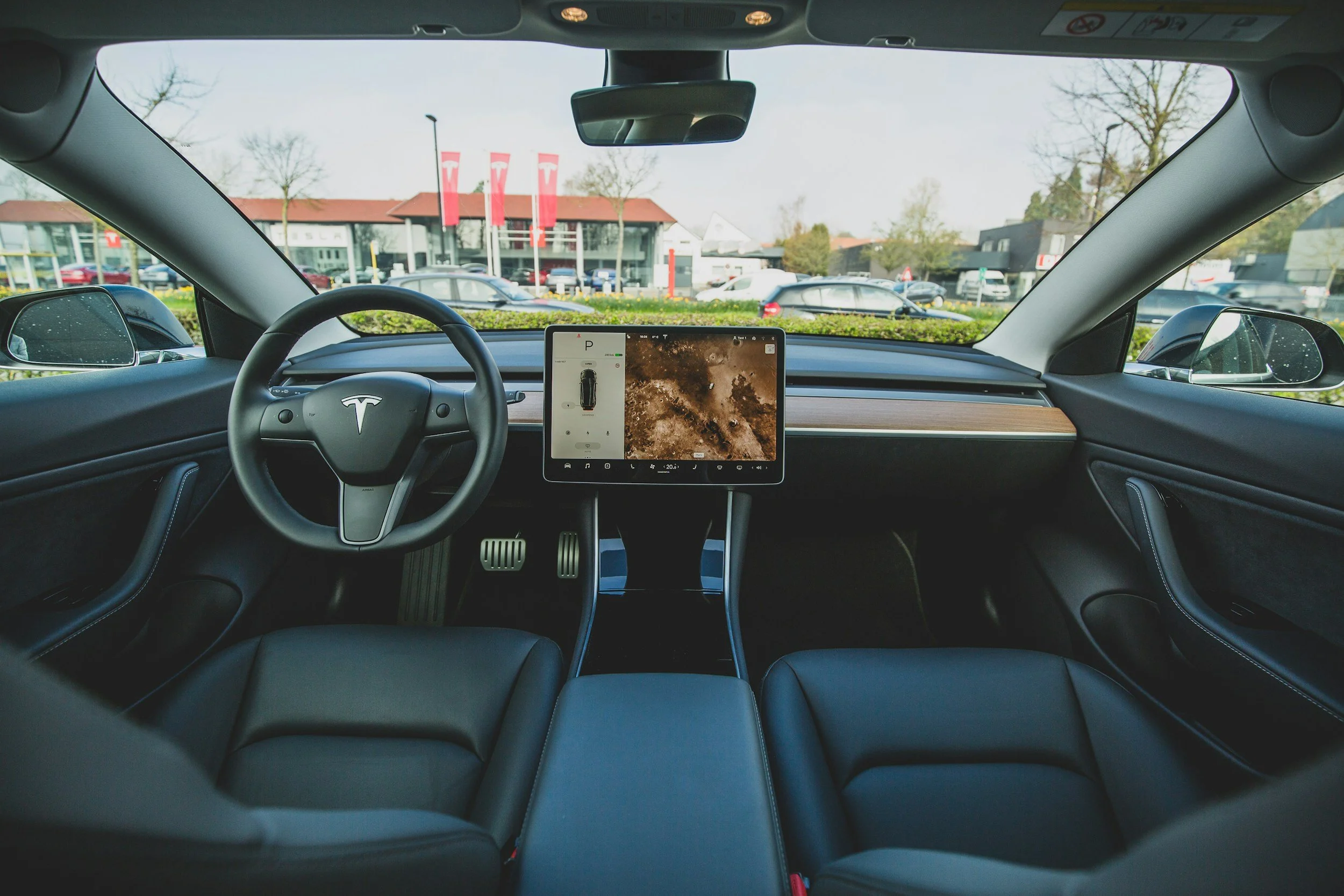 Inside a Tesla electric vehicle, showing the front interior with a steering wheel, dashboard, and large central touchscreen display, parked in a lot with a Tesla store and cars visible outside.