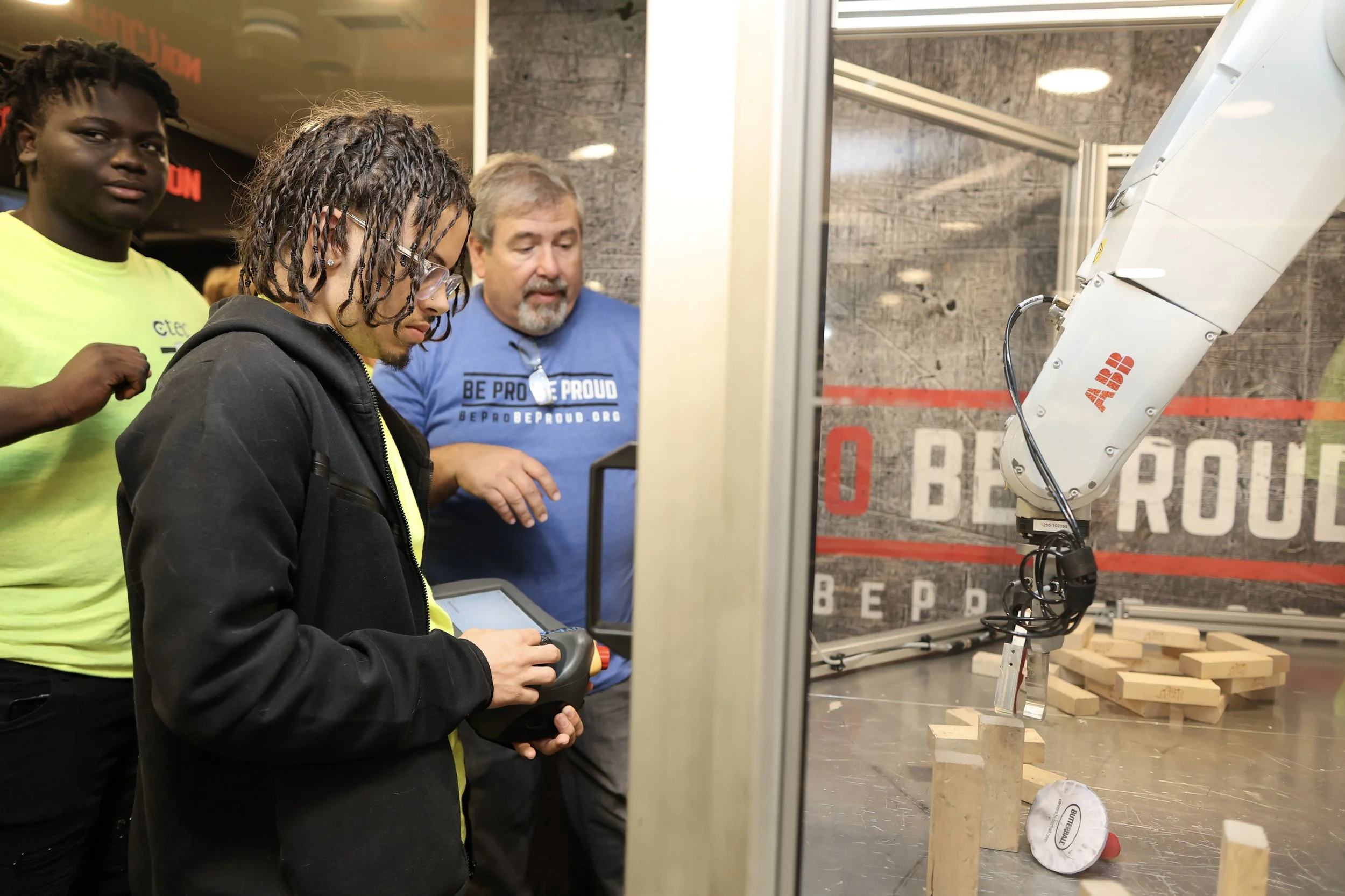 Three people observing a mechanical arm in an indoor exhibition. One man is wearing a blue shirt, and two young men are wearing black and yellow shirts. The robotic arm is grasping a wooden block, and there are more wooden blocks on the table.