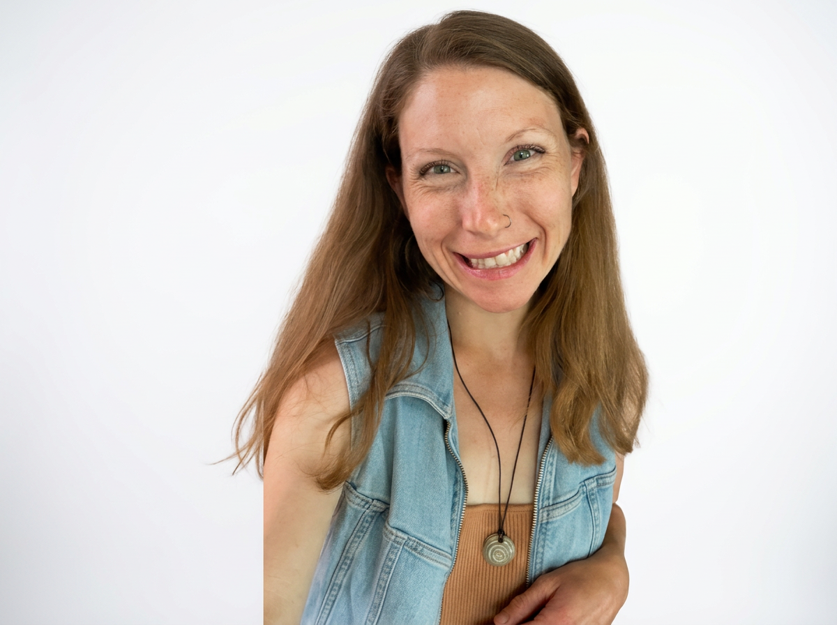 A smiling woman with long red hair, freckles, and a nose piercing, wearing a denim vest over a brown top and a necklace with a spiral pendant, standing against a plain white background.