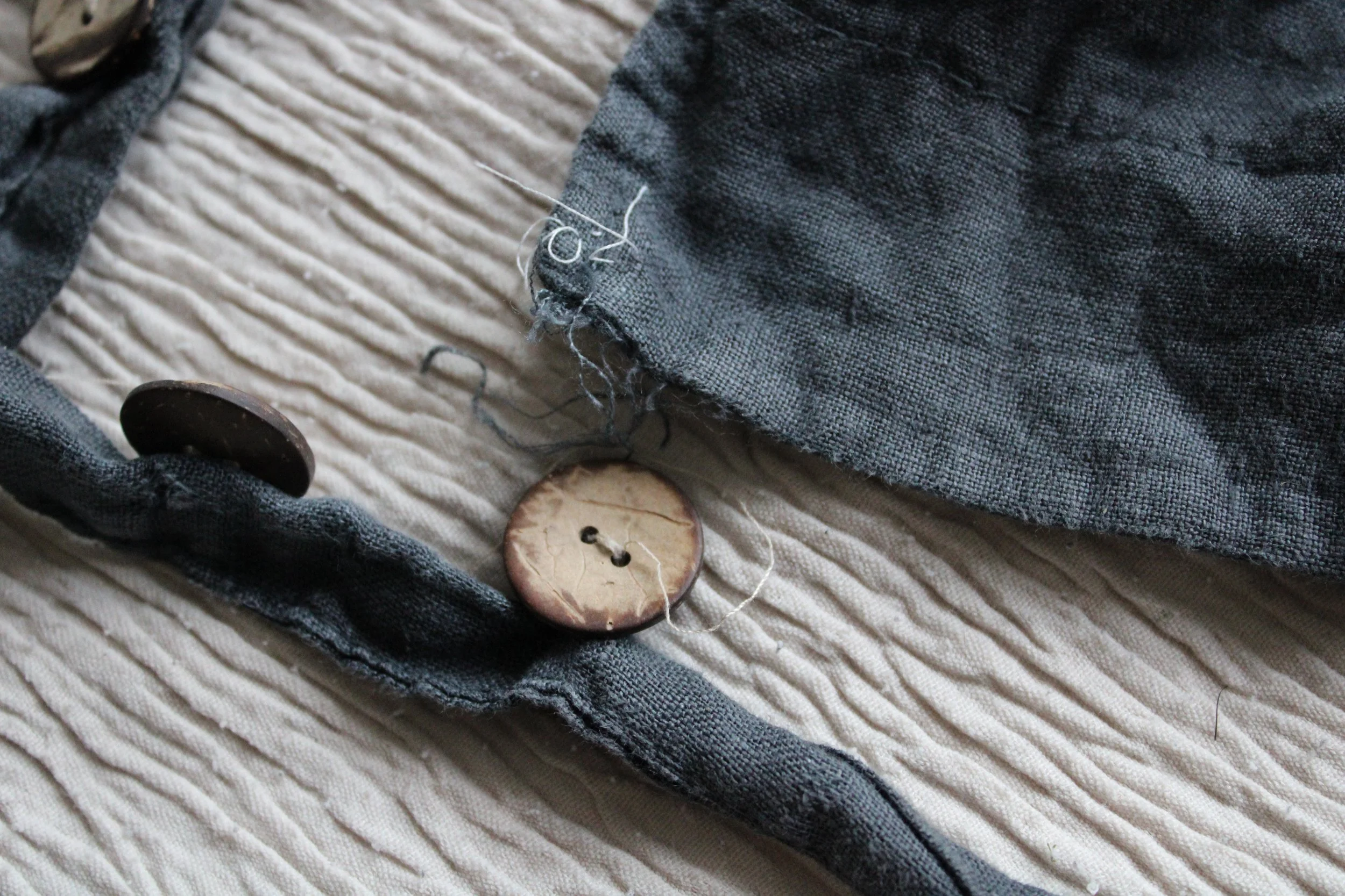Close-up of a worn-out blue jeans fabric with a small frayed tear near the seam, resting on a beige textured fabric surface, with a large brown button nearby.