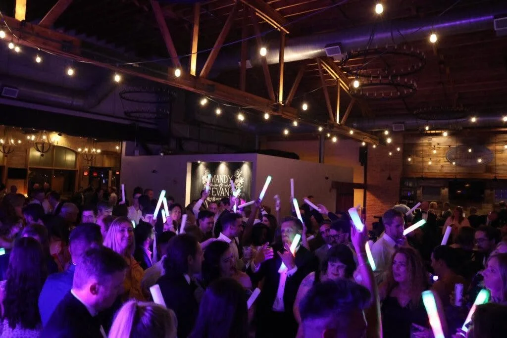 Crowd of people at a party or concert holding glowing light sticks, in a dimly lit venue with string lights and a dance floor.