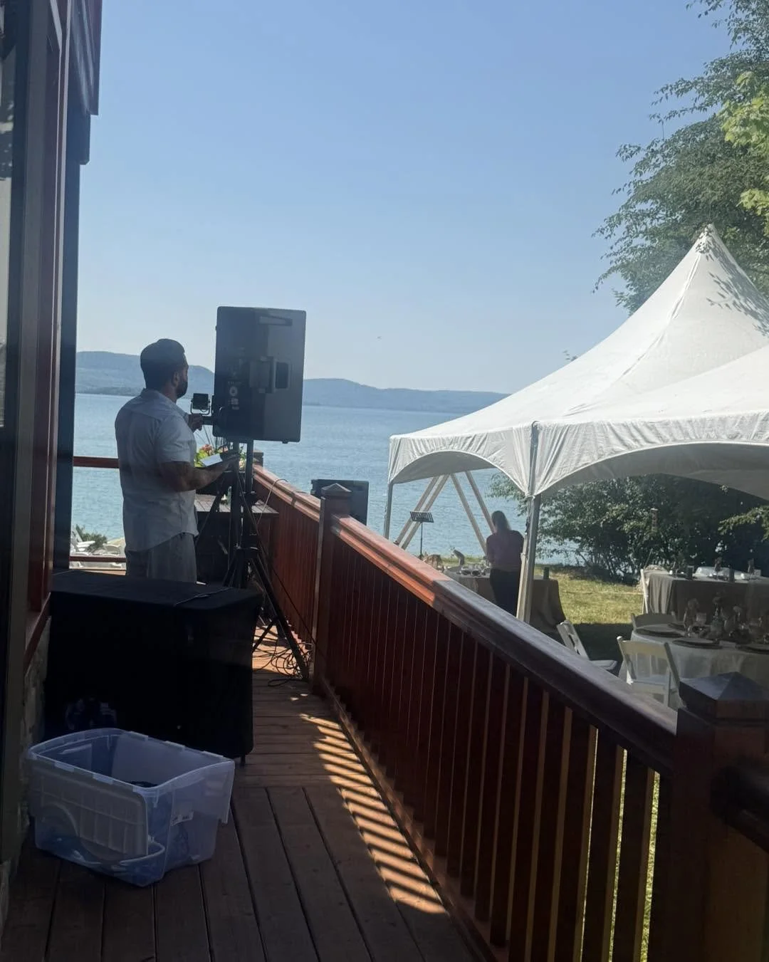 A man setting up audiovisual equipment on a wooden deck overlooking a lake with mountains in the distance. There is a white canopy and chairs set up for an outdoor event.