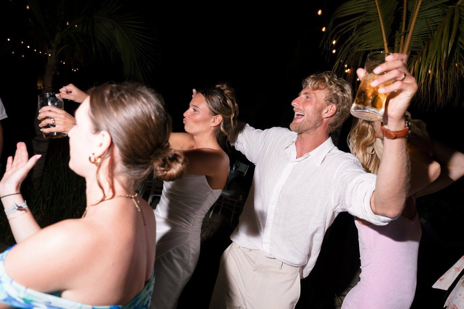 People enjoying a lively party outdoors at night, dancing and holding drinks, with string lights and palm trees in the background.