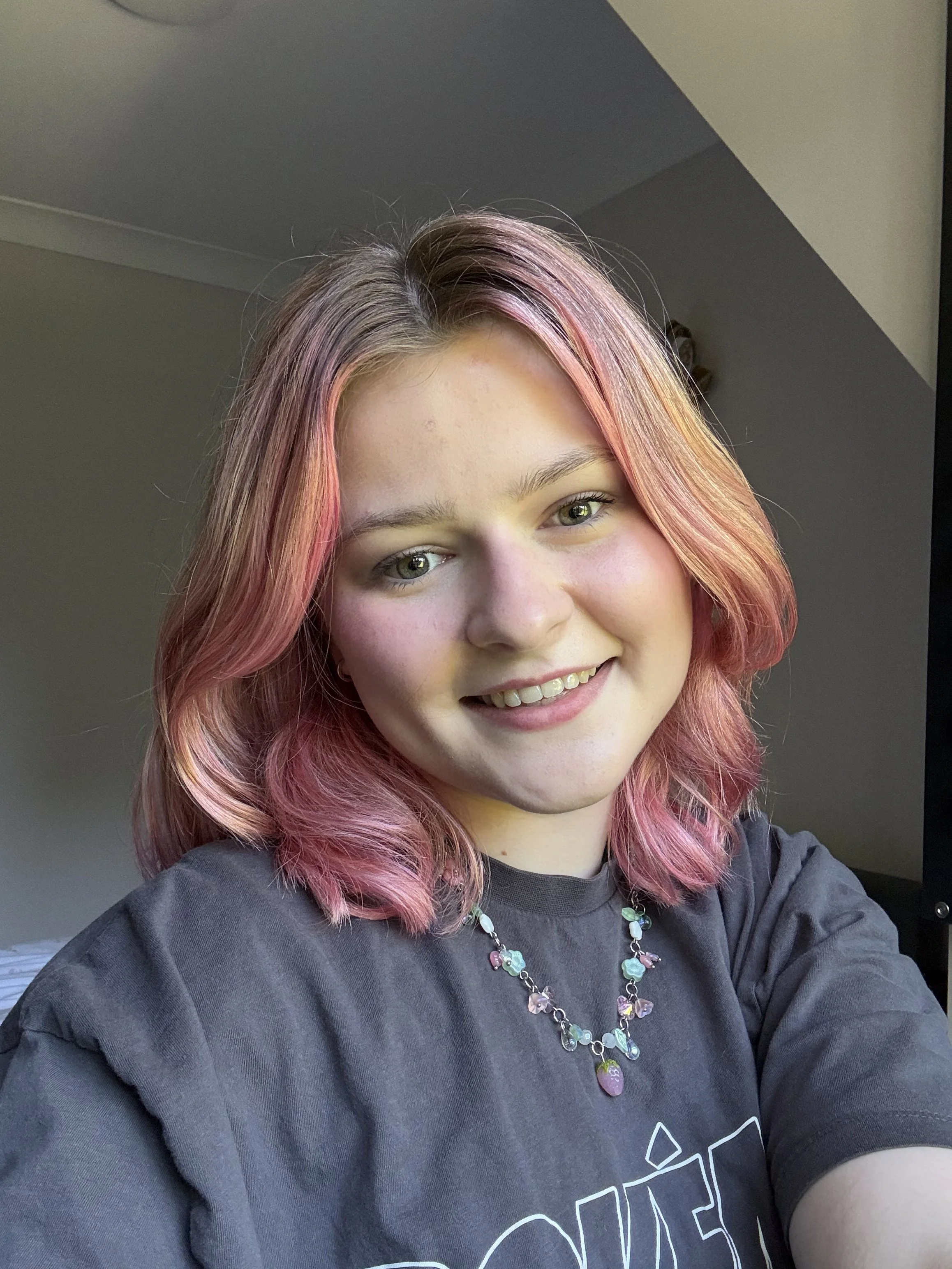 Author photo of Mia R Donovan, a young woman in her early twenties with pink shoulder-length hair, hazel eyes and fair eyebrows. She smiles at the camera and wears a grey t-shirt with a Strawberry Shortcake glass necklace from MarsNStars Jewellery.