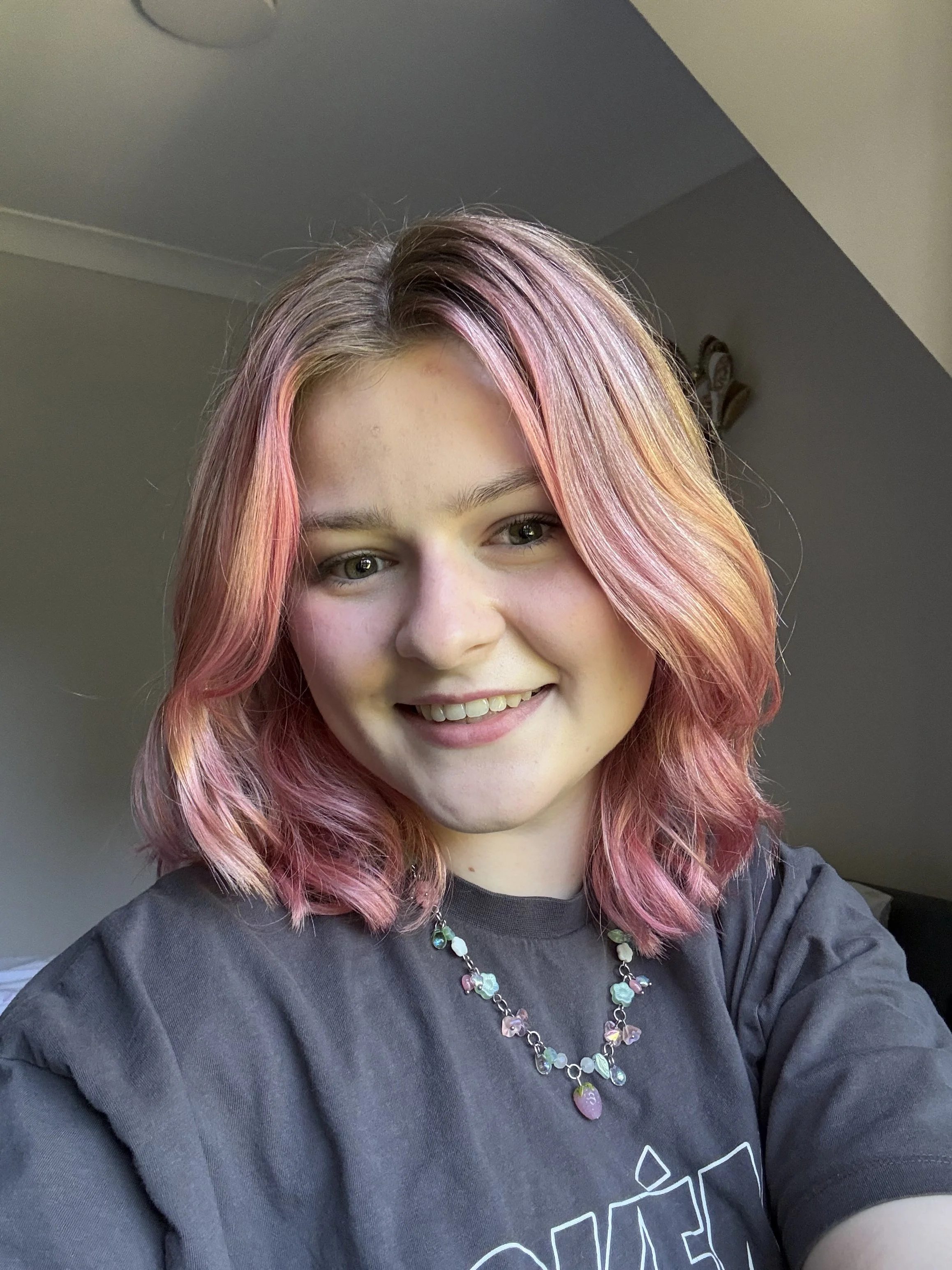 Author photo of Mia R Donovan, a young woman in her early twenties with pink shoulder-length hair, hazel eyes and fair eyebrows. She smiles at the camera and wears a grey t-shirt with a Strawberry Shortcake glass necklace from MarsNStars Jewellery.