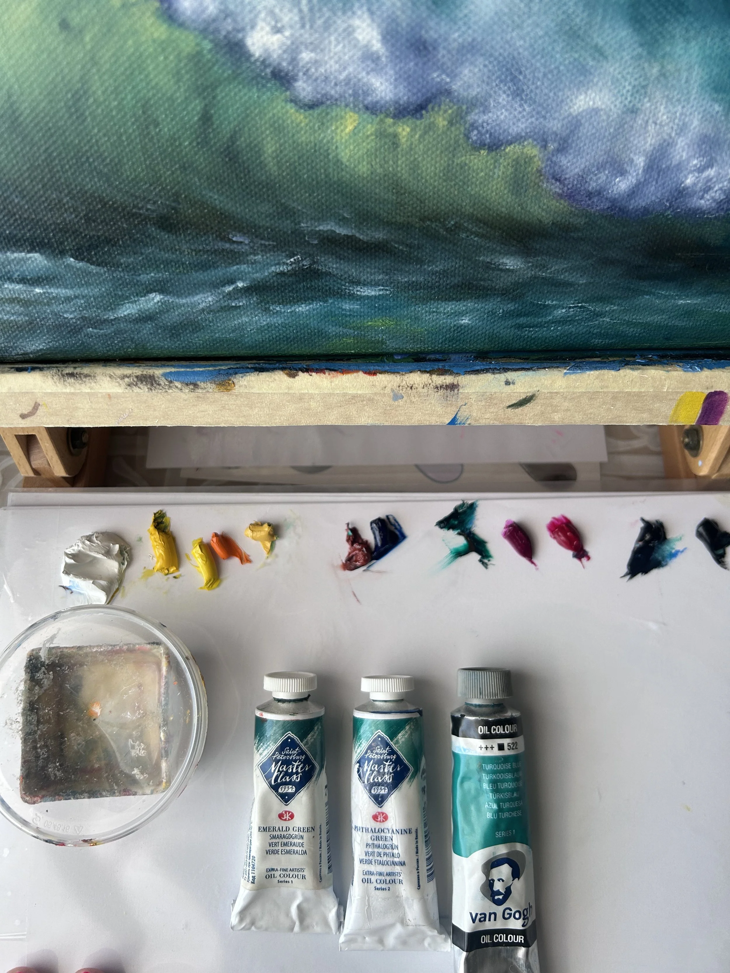 Set of three oil paint tubes labeled 'Master Class' in emerald green, phthalocyanine green, and van Gogh turquoise, placed on a white surface with spatters of paint, alongside a palette with mixed colors, a container of water, and a shelf with paint on it.