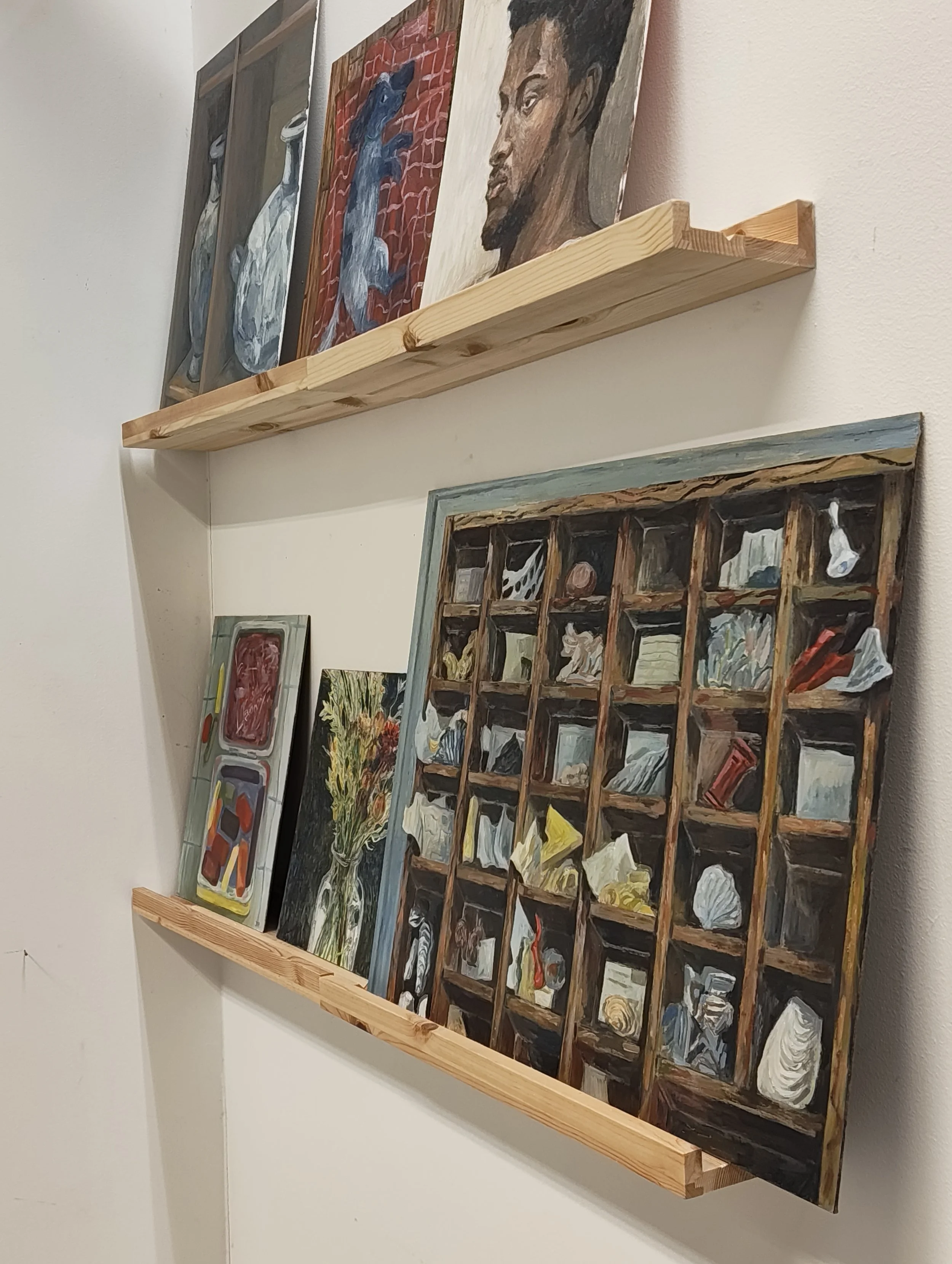 Four paintings on a wall, with two displayed on a wooden shelf. The paintings depict various subjects, including a vase with yellow flowers, a character with a dark background, and a detailed grid-style composition with multiple smaller images. The paintings are framed and leaning against the wall.