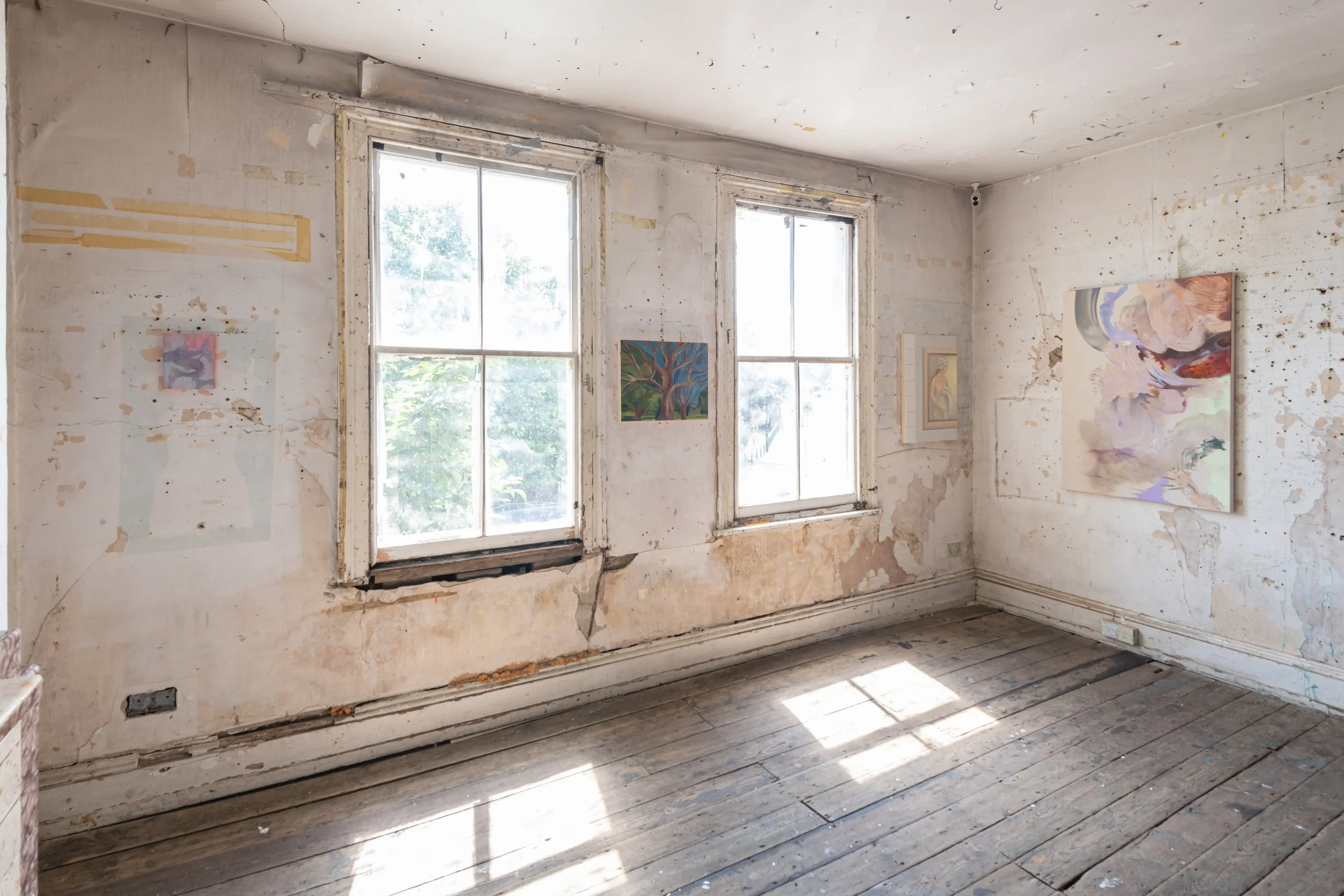 An empty, unfinished room with peeling paint, exposed wood floorboards, and large windows allowing sunlight to brighten the space; several small, colorful paintings are hung on the wall.