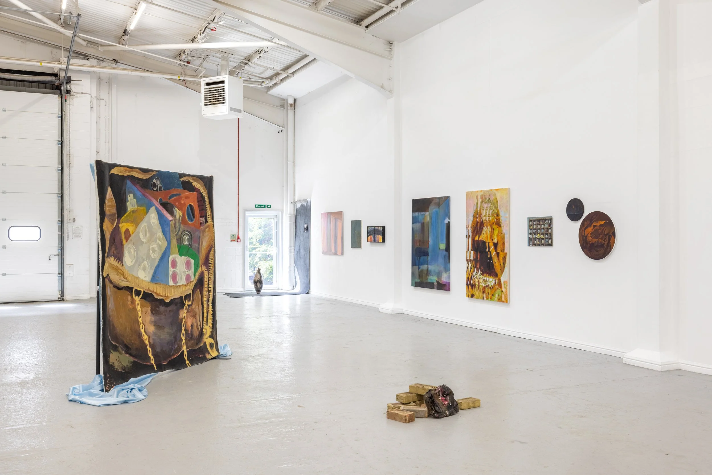 An art gallery with white walls and various colorful paintings displayed. In the foreground, a large abstract tapestry with chains and gold accents is draped over a stand. On the floor, there are bricks and a small piece of artwork.