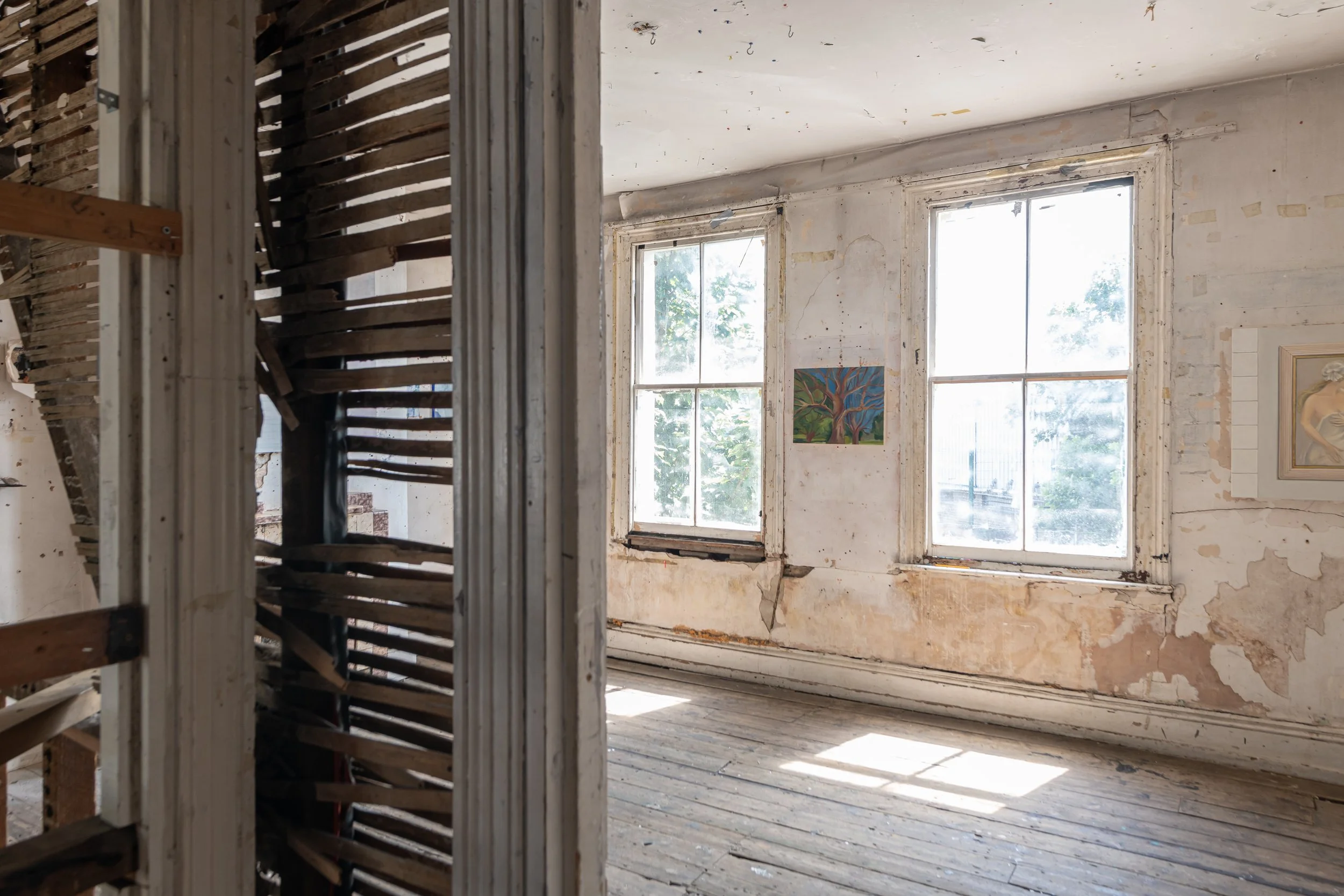View of an interior room with peeling paint on walls, old windows, and a wooden floor, seen through a partially dismantled staircase.