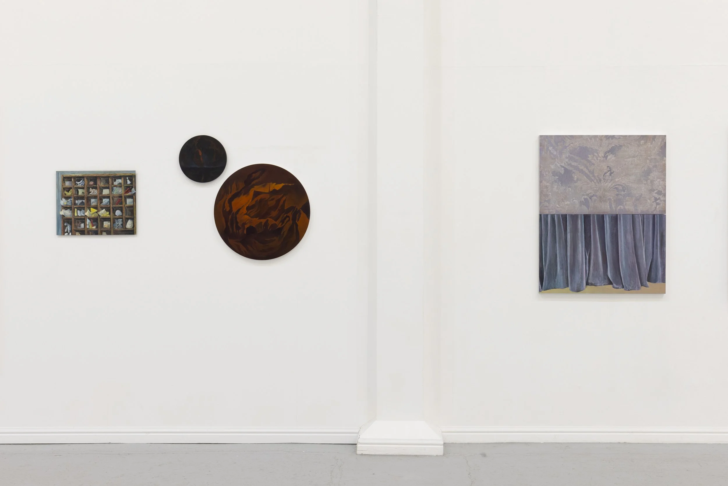 A gallery wall with five pieces of artwork: a small framed collage of various objects on the far left, two round dark-colored abstract paintings in the middle, and a large rectangular piece on the right that appears to be an abstract painting with a 