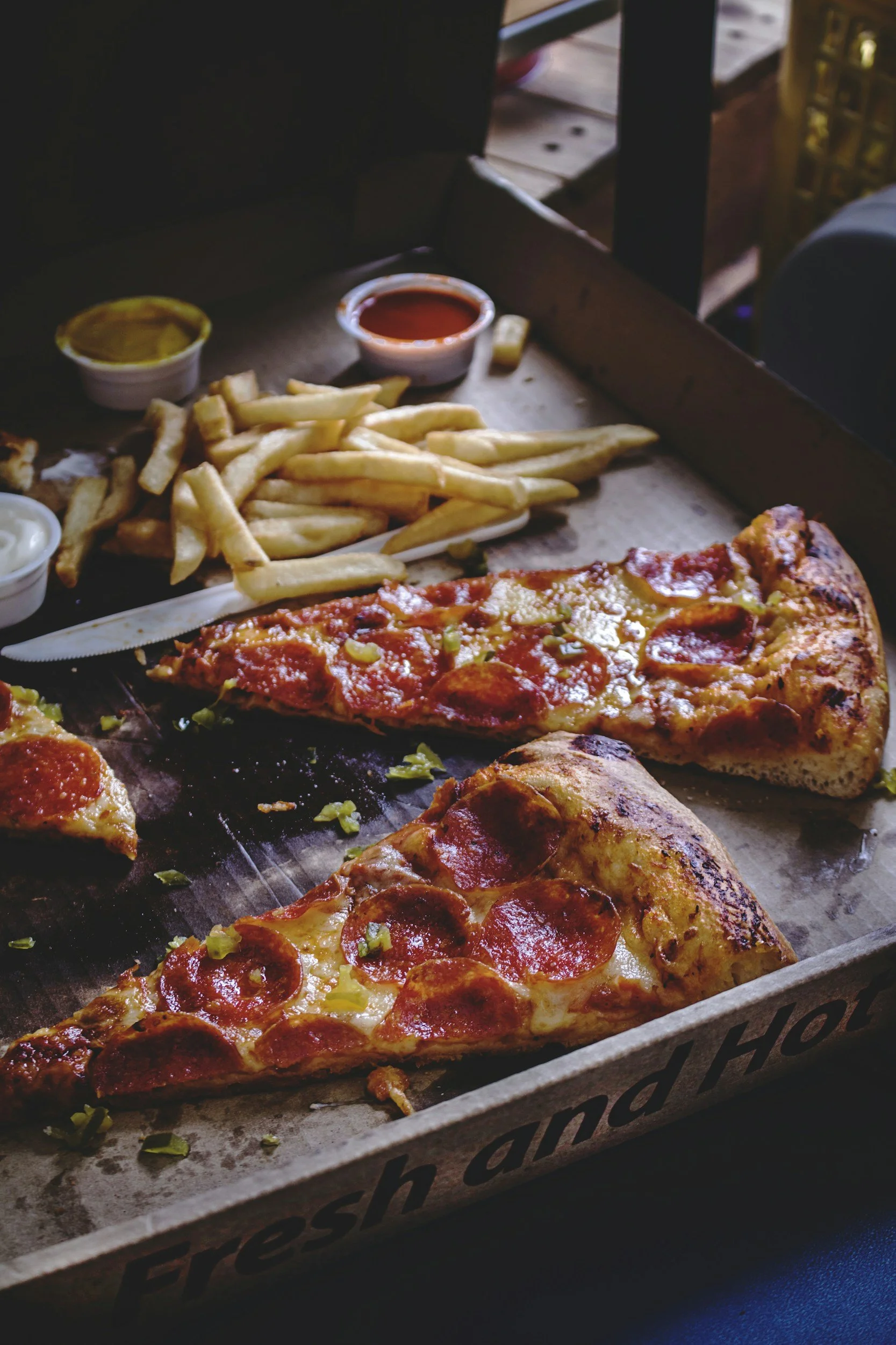 Box of pizza slices with pepperoni and cheese, French fries, and small containers of ketchup and mustard inside a pizza box.