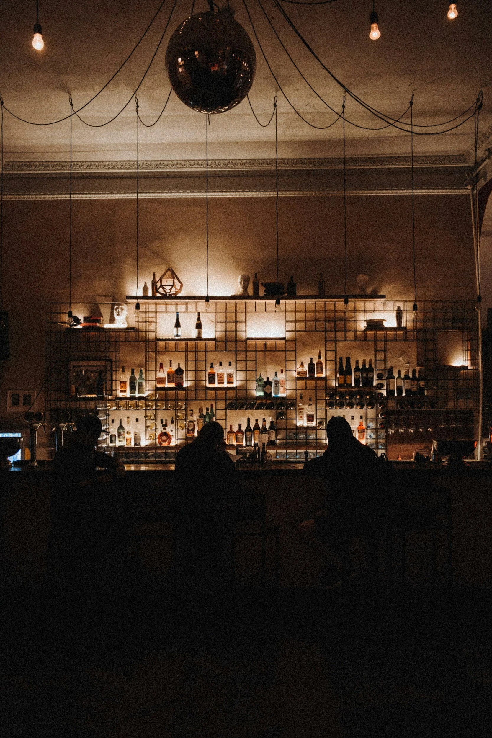Dimly lit bar with a mirrored disco ball hanging from the ceiling, string lights, and a shelf with bottles of alcohol and decorative objects, with patrons sitting at the bar in silhouette.