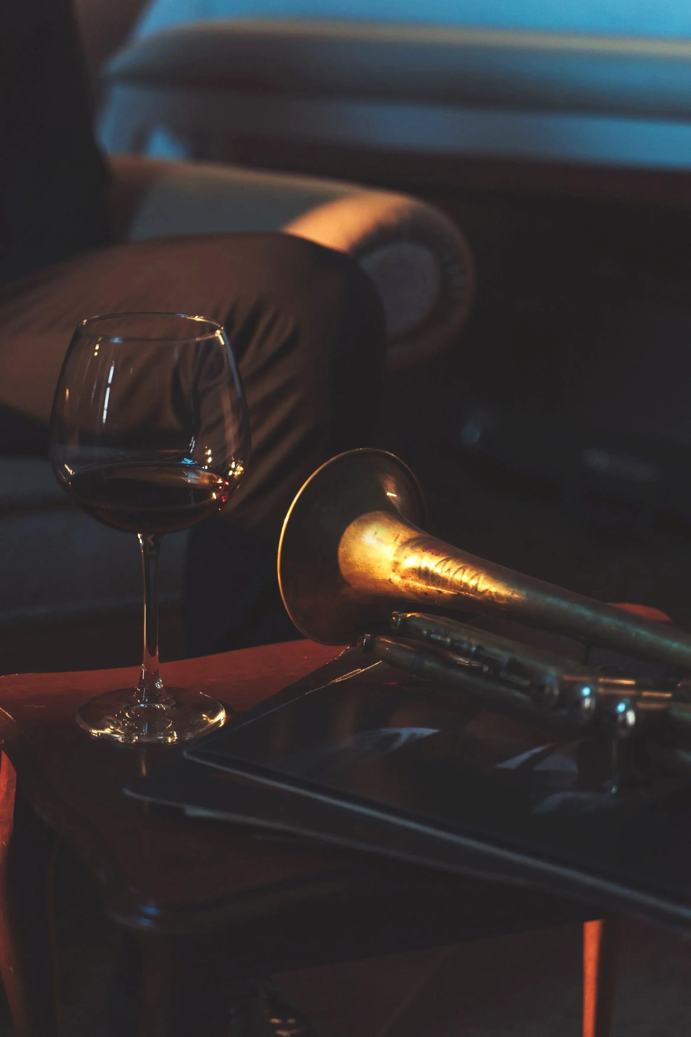 A glass of red wine on a table with a wine opener and a tray, with a blurred background of a person reclining and a boomerang-style lamp.