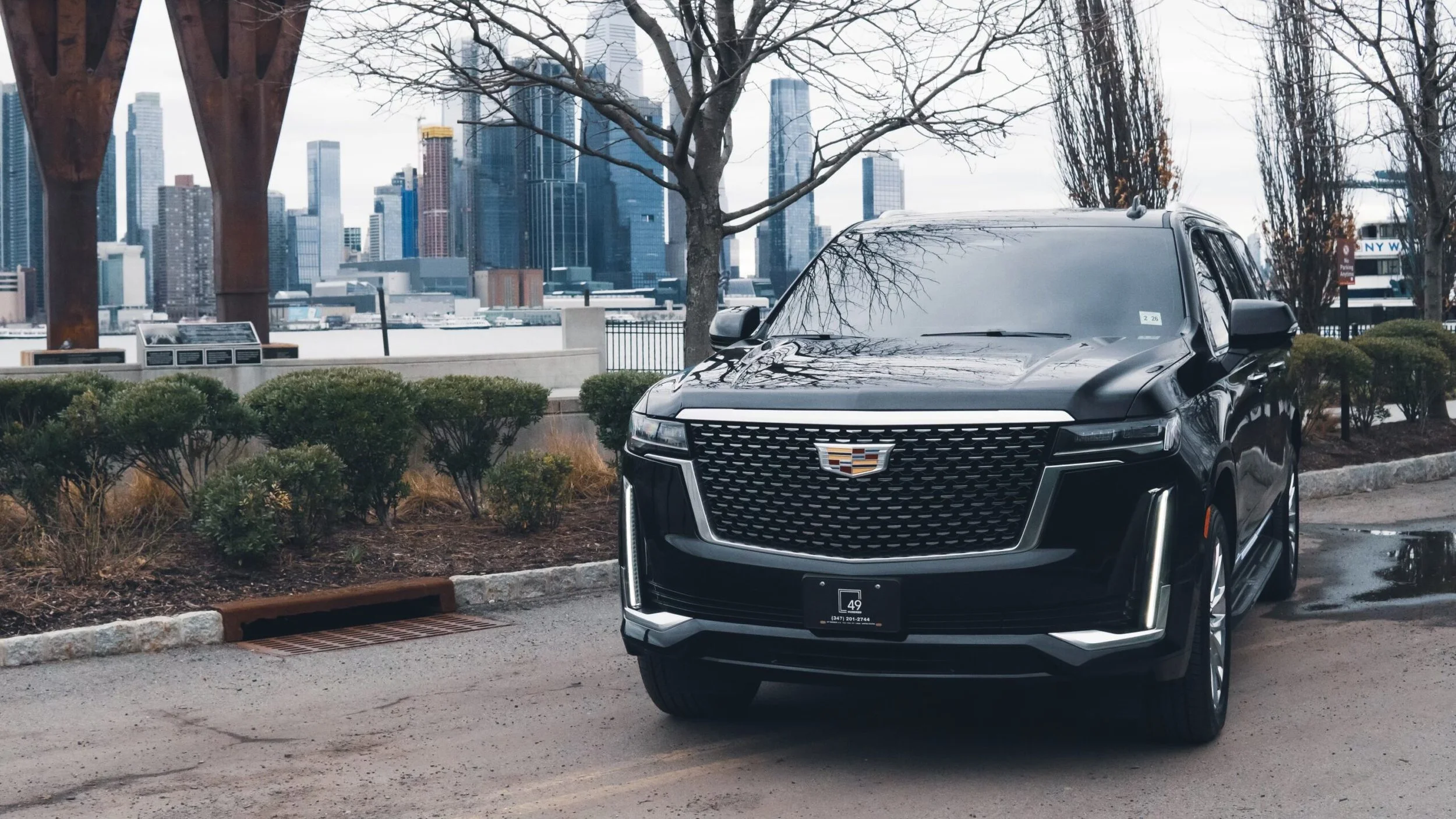 A black Cadillac SUV parked near a sidewalk with city skyscrapers in the background on a cloudy day.