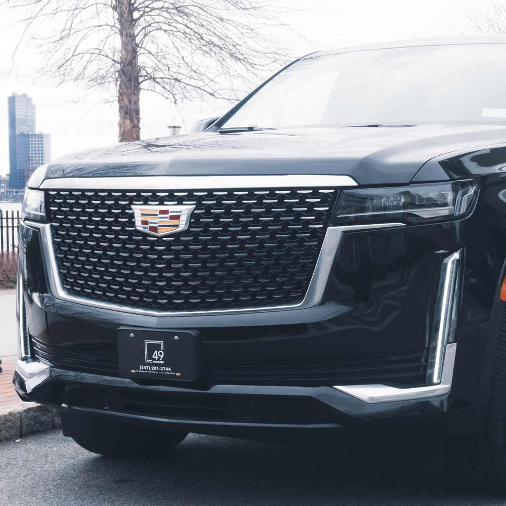 Close-up of the front grille of a black Cadillac vehicle, showing the Cadillac emblem and part of the windshield with a cityscape in the background.