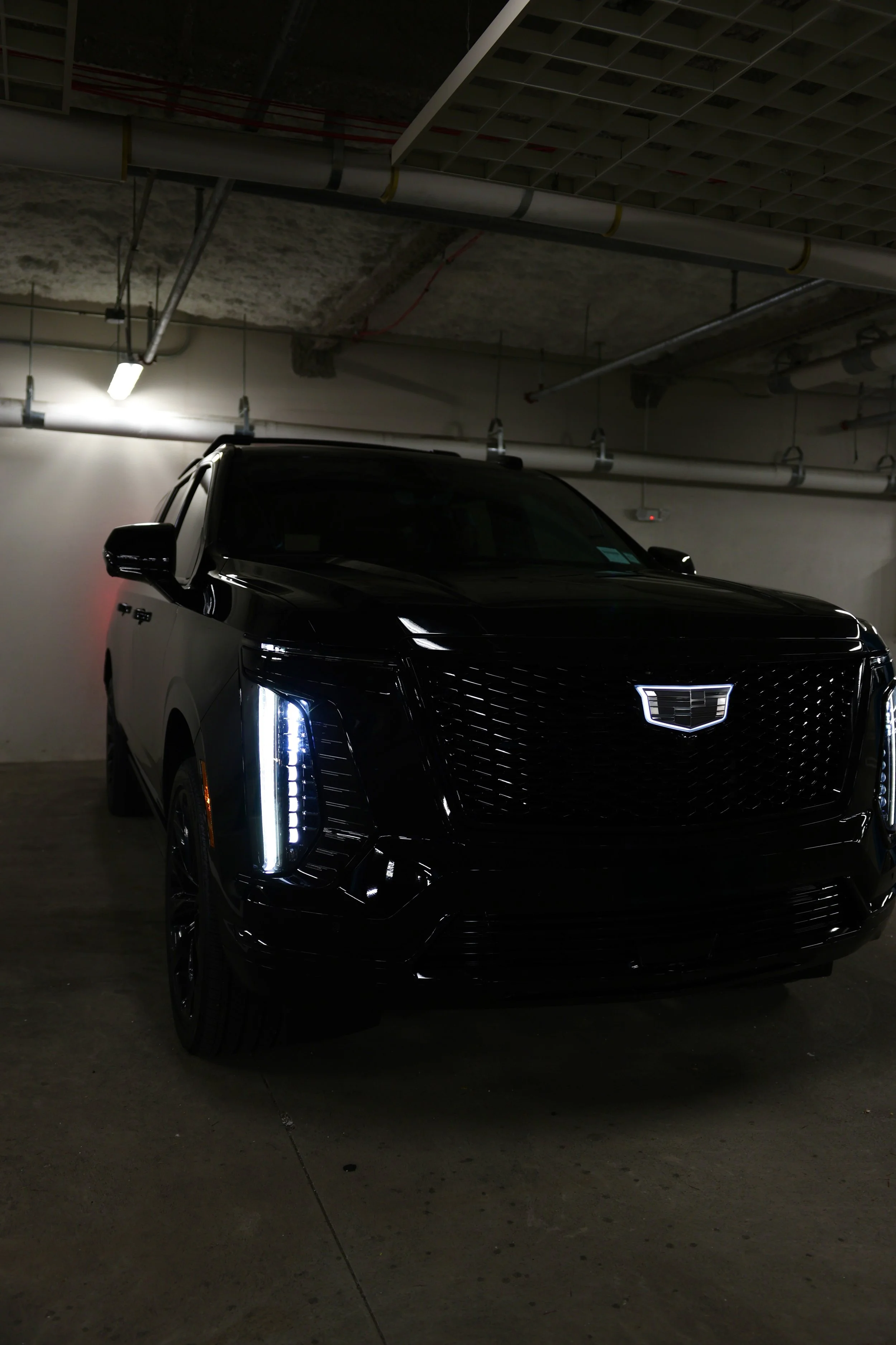 A black Cadillac SUV parked in an underground parking garage with visible pipes and lighting overhead.