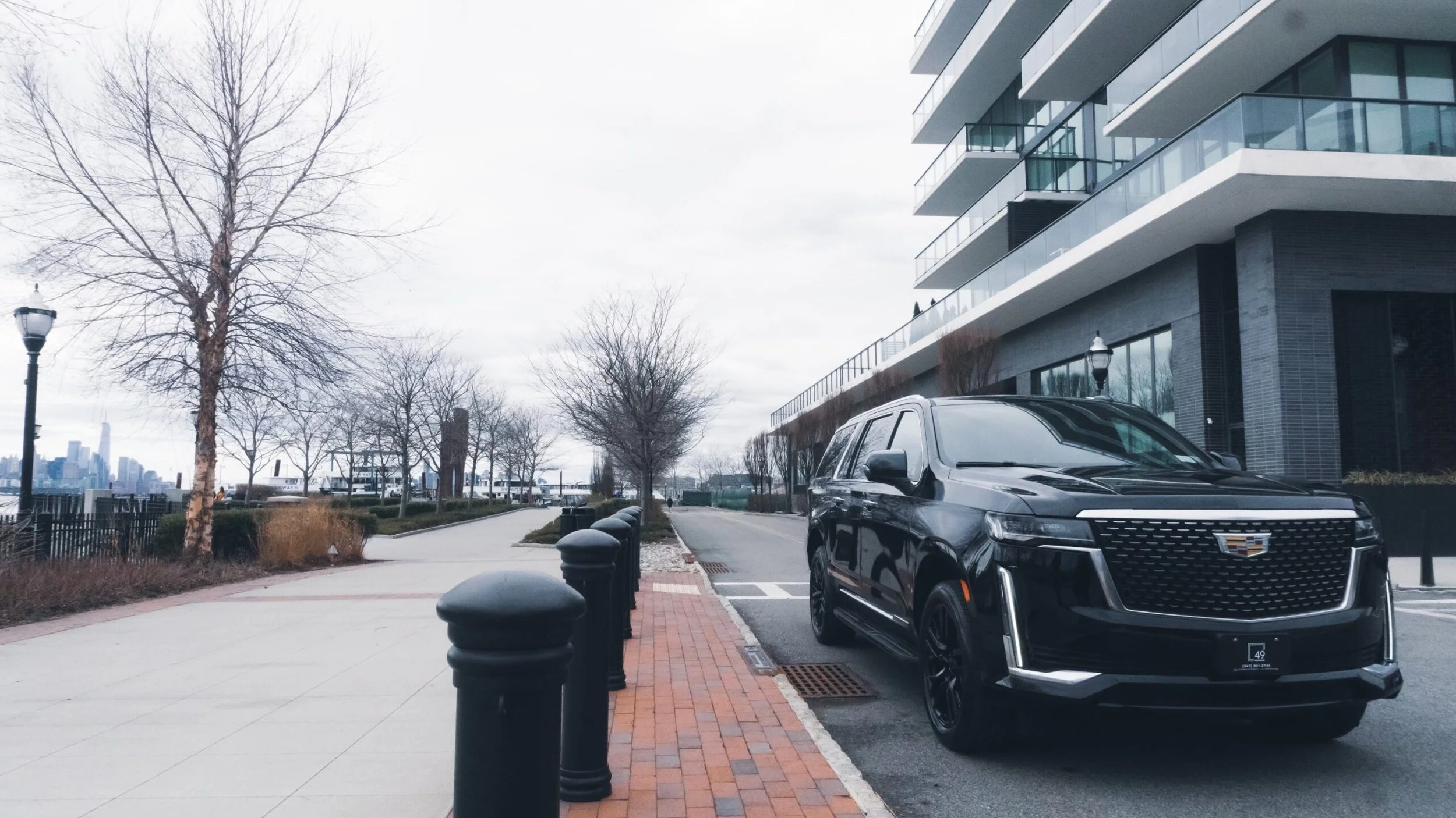 A modern black Cadillac SUV parked on a city street with a sidewalk, leafless trees, and a multi-story residential building with glass balconies, overcast sky, and a distant city skyline.