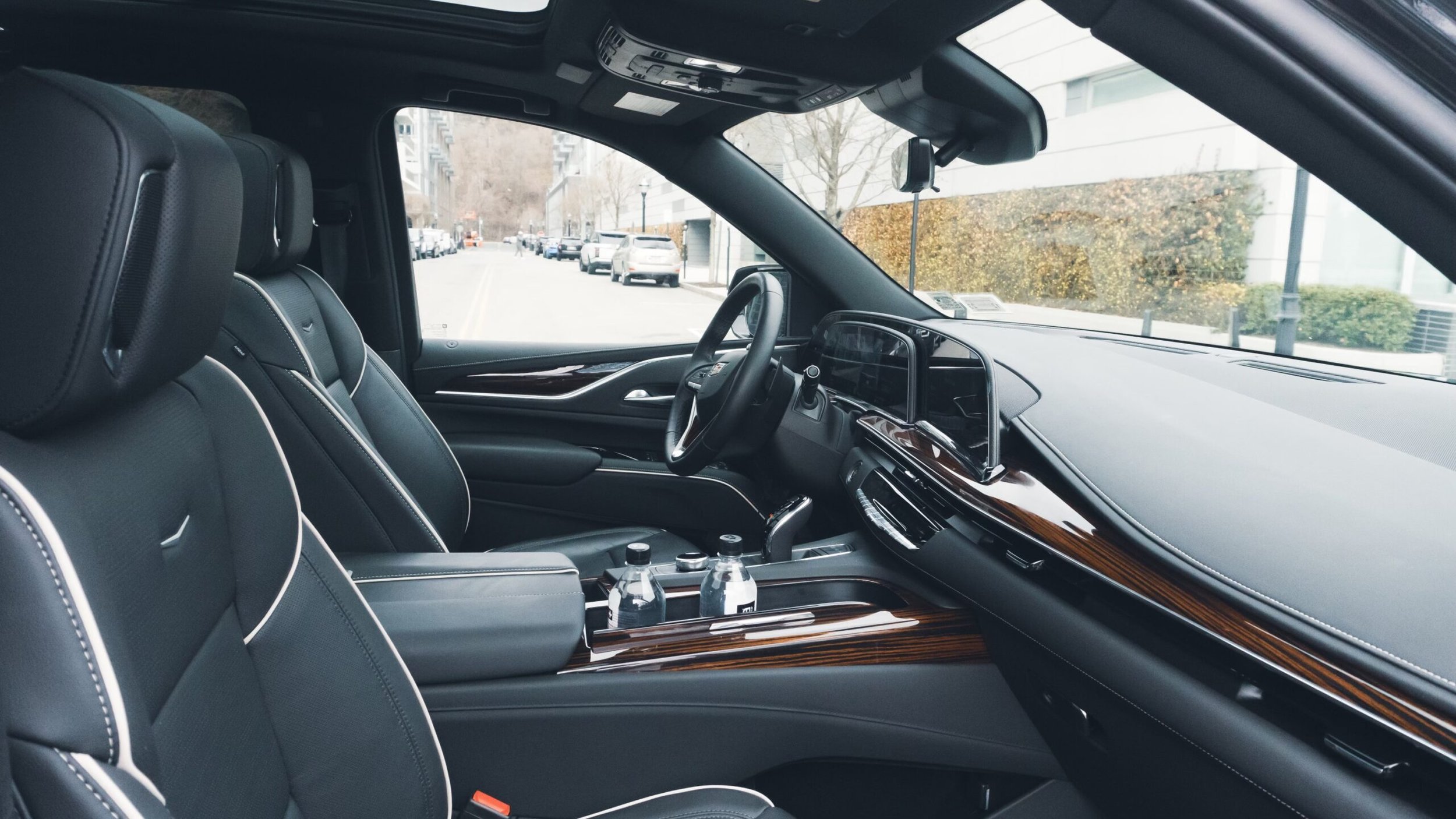 Interior of a luxury vehicle showing black leather seats with white stitching, a dashboard with a touchscreen display, and two water bottles in the center console, with a view of an urban street outside.