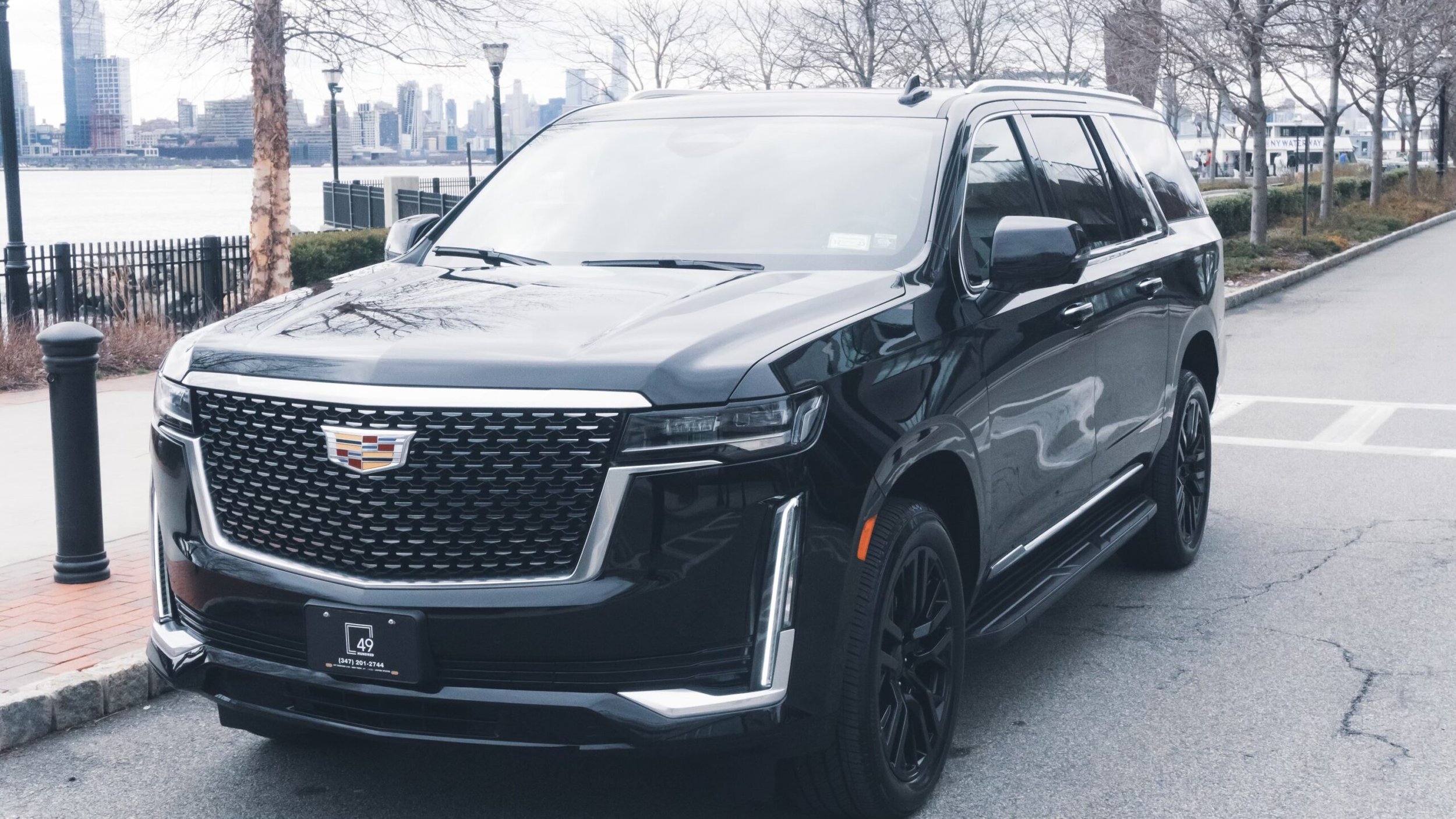 A black Cadillac SUV parked on a city street near a waterfront with a city skyline in the background. There are trees, lampposts, and a railing along the water.