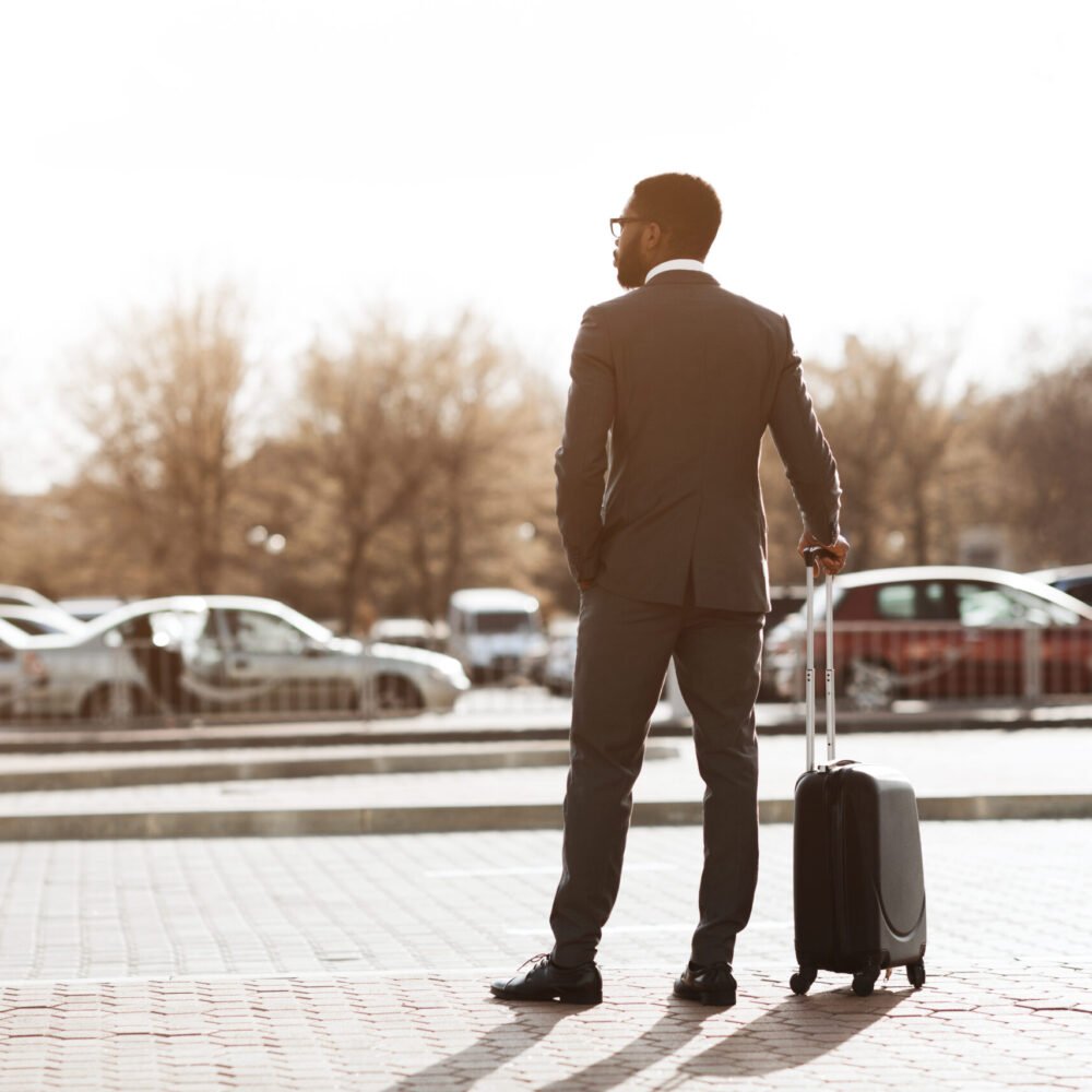 A man in a suit with glasses standing outdoors, holding a rolling suitcase, with parked cars and leafless trees in the background.
