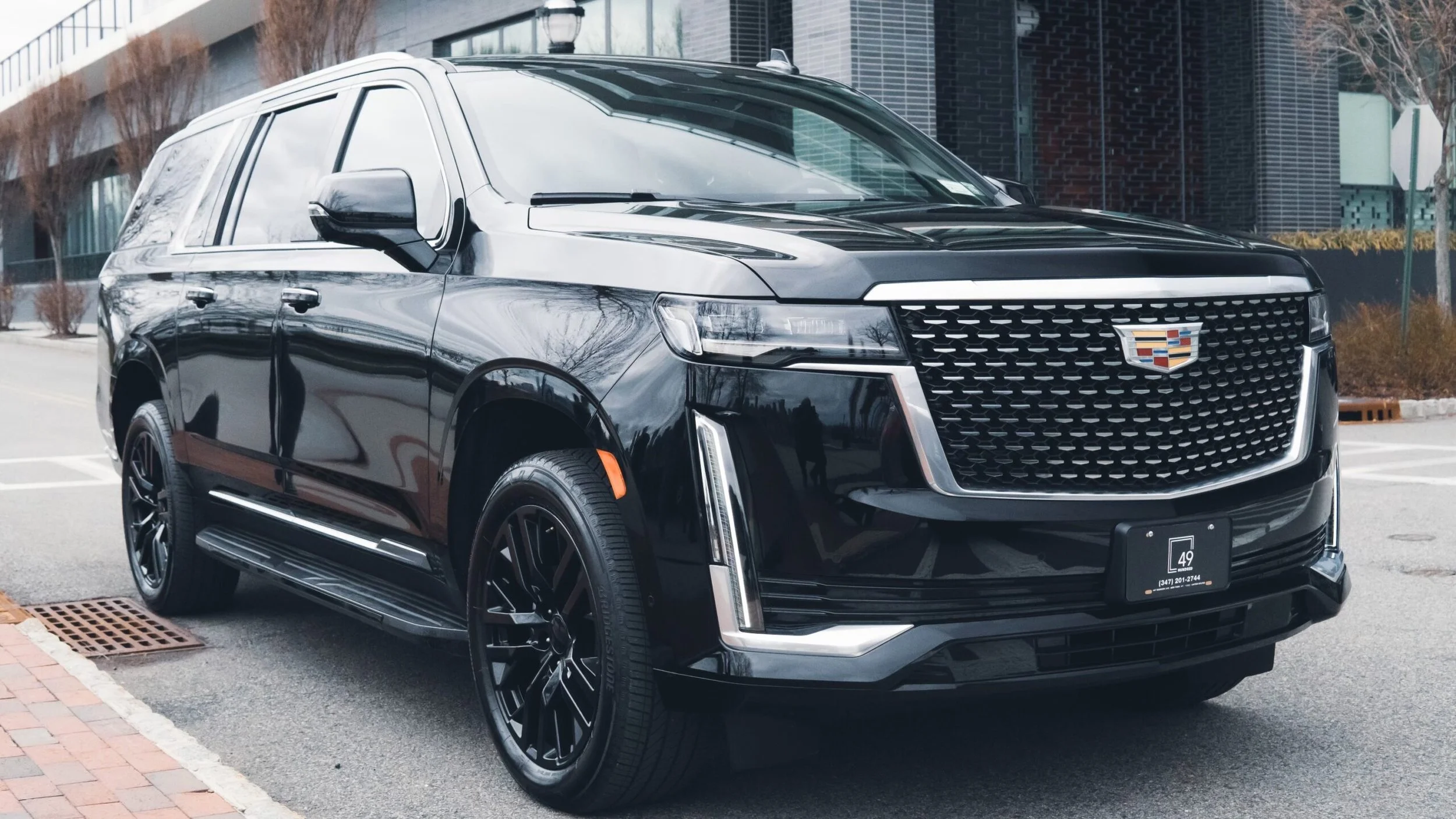 Black Cadillac SUV parked on the street in front of a modern building, with trees and a sidewalk nearby.