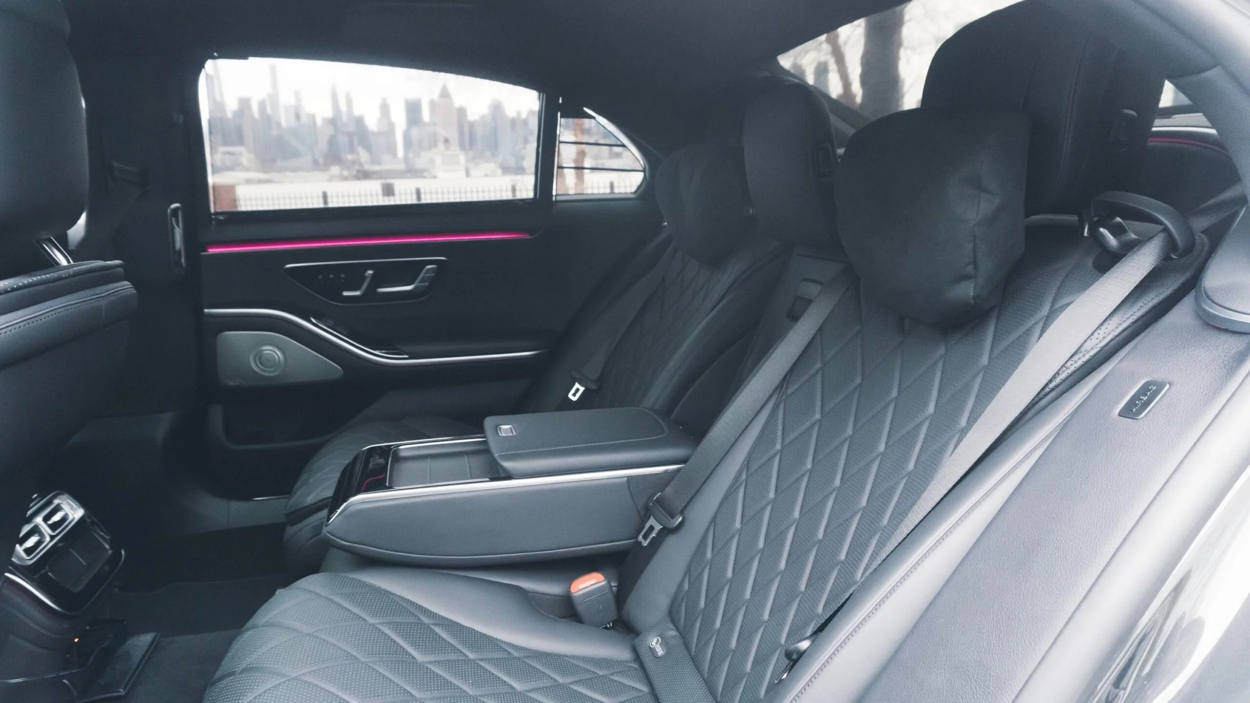 Interior of a luxury car showing black leather seats with quilted stitching, headrests, and a city skyline visible through the window.
