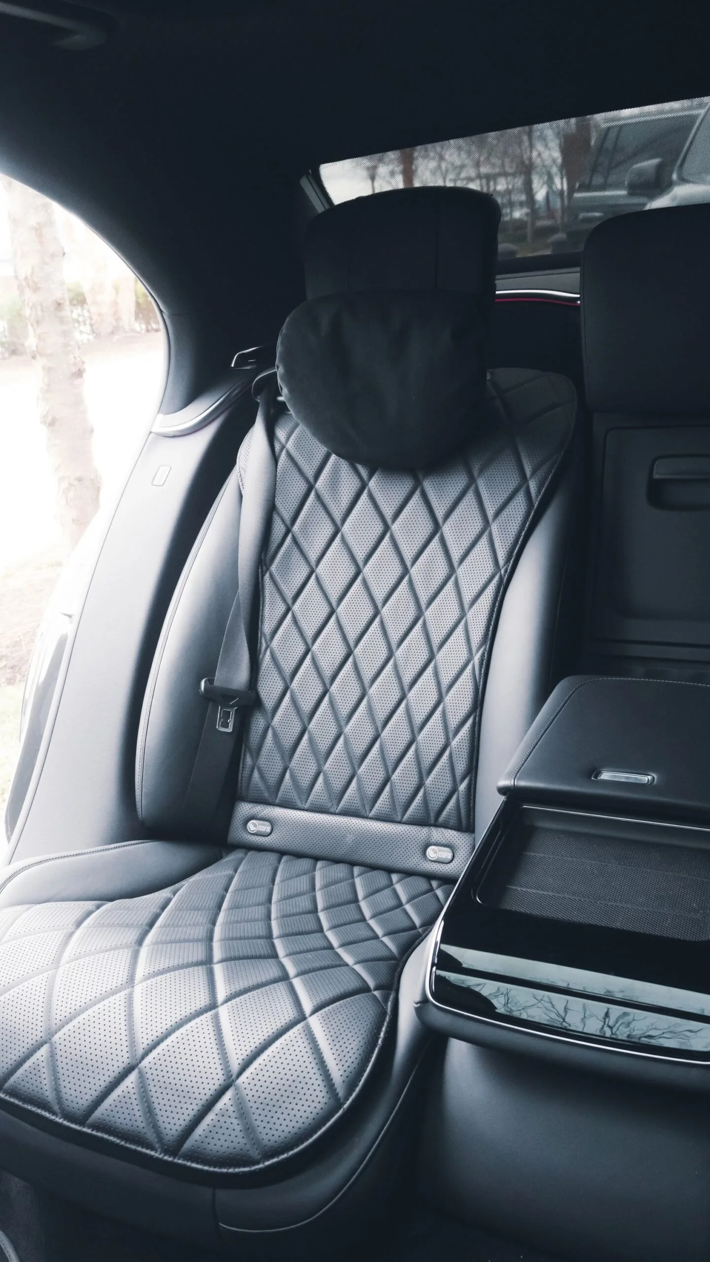 The image shows a luxurious car seat with quilted black and gray upholstery, a headrest with a black cover, a seatbelt, a window with a view of trees outside, and a side storage compartment.