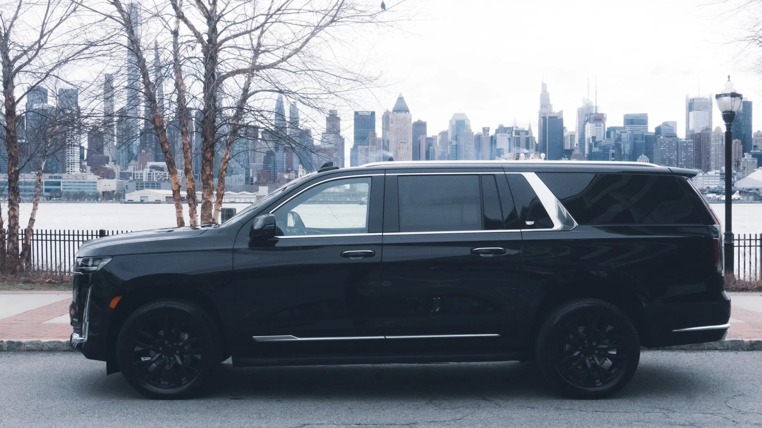 Black SUV parked near tree-lined waterfront with city skyline in the background.