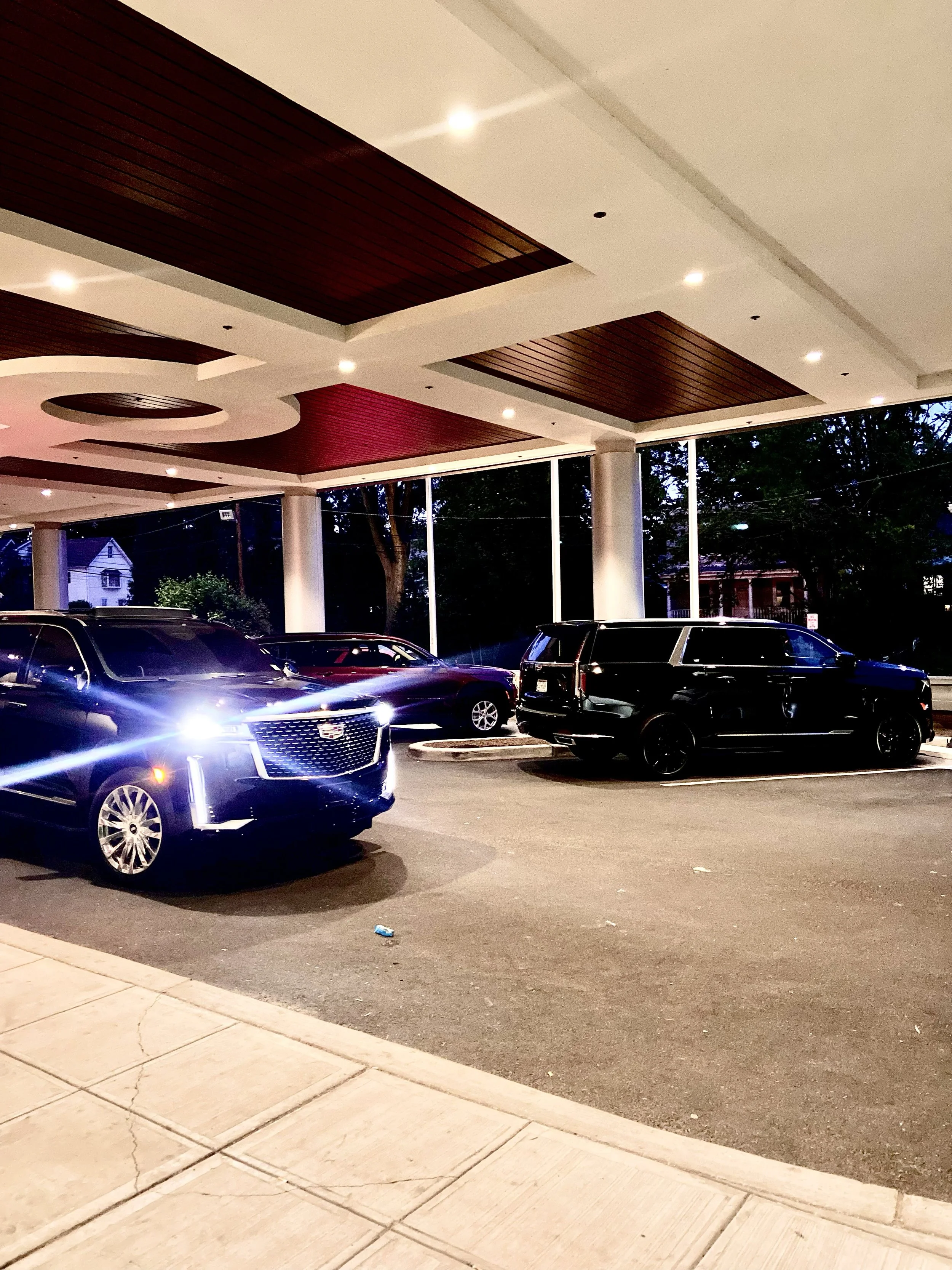 Nighttime scene of a parking lot with several parked cars underneath a modern architectural canopy featuring wood paneling and embedded lights. Trees and residential houses are visible in the background.