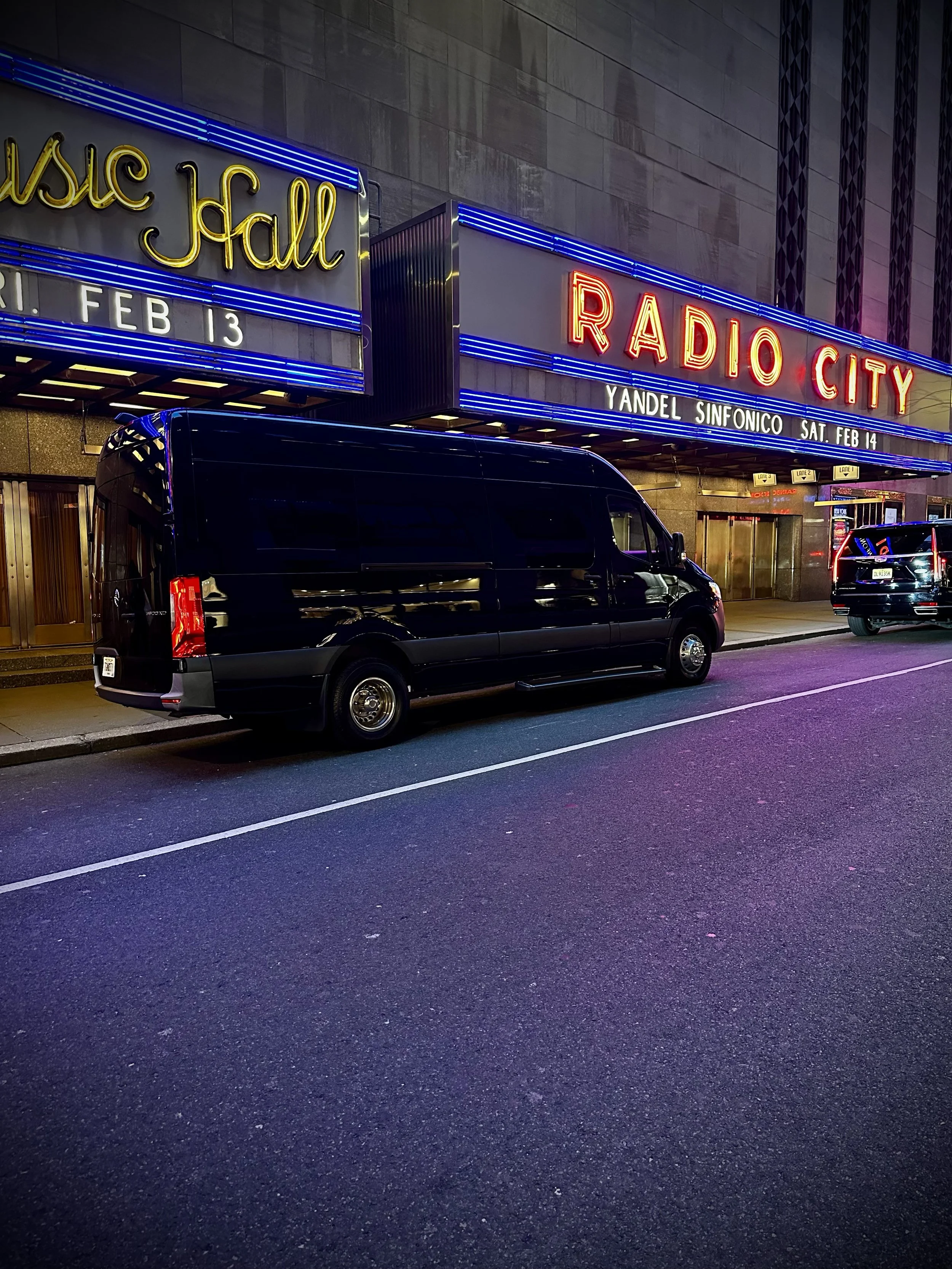 Night view of the front of a theater marquee for Radio City Music Hall, with neon lights displaying the date February 13 and an upcoming concert by Yandel Sinfonico on Saturday, February 14. Vehicles are parked in front of the theater.