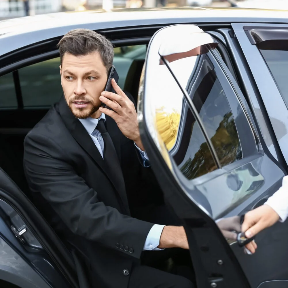 A man in a suit sitting inside a car, talking on a cell phone, with someone handing him car keys through the open car door.