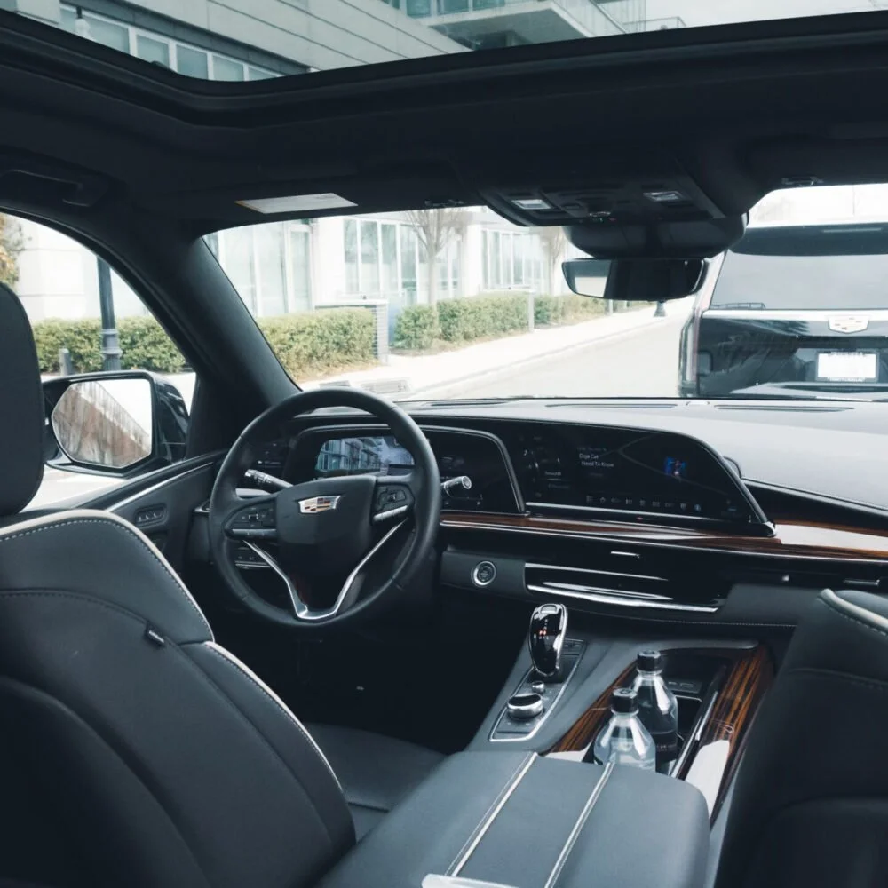 Interior view of a Cadillac SUV showing the steering wheel, dashboard, center console with gear shifter, and two water bottles, with a parking lot and buildings visible through the windshield.