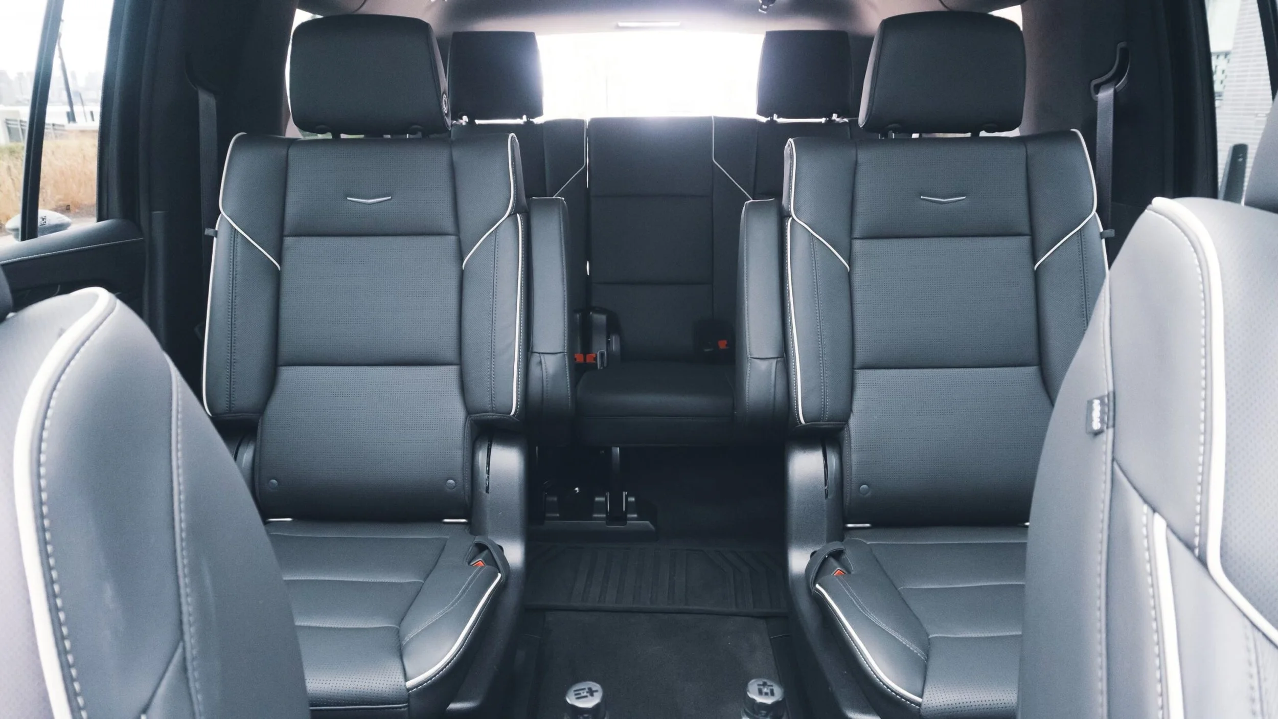 The interior of a vehicle showing three rows of black leather seats with white trim, black headrests, and a view through the front windshield of buildings outside.