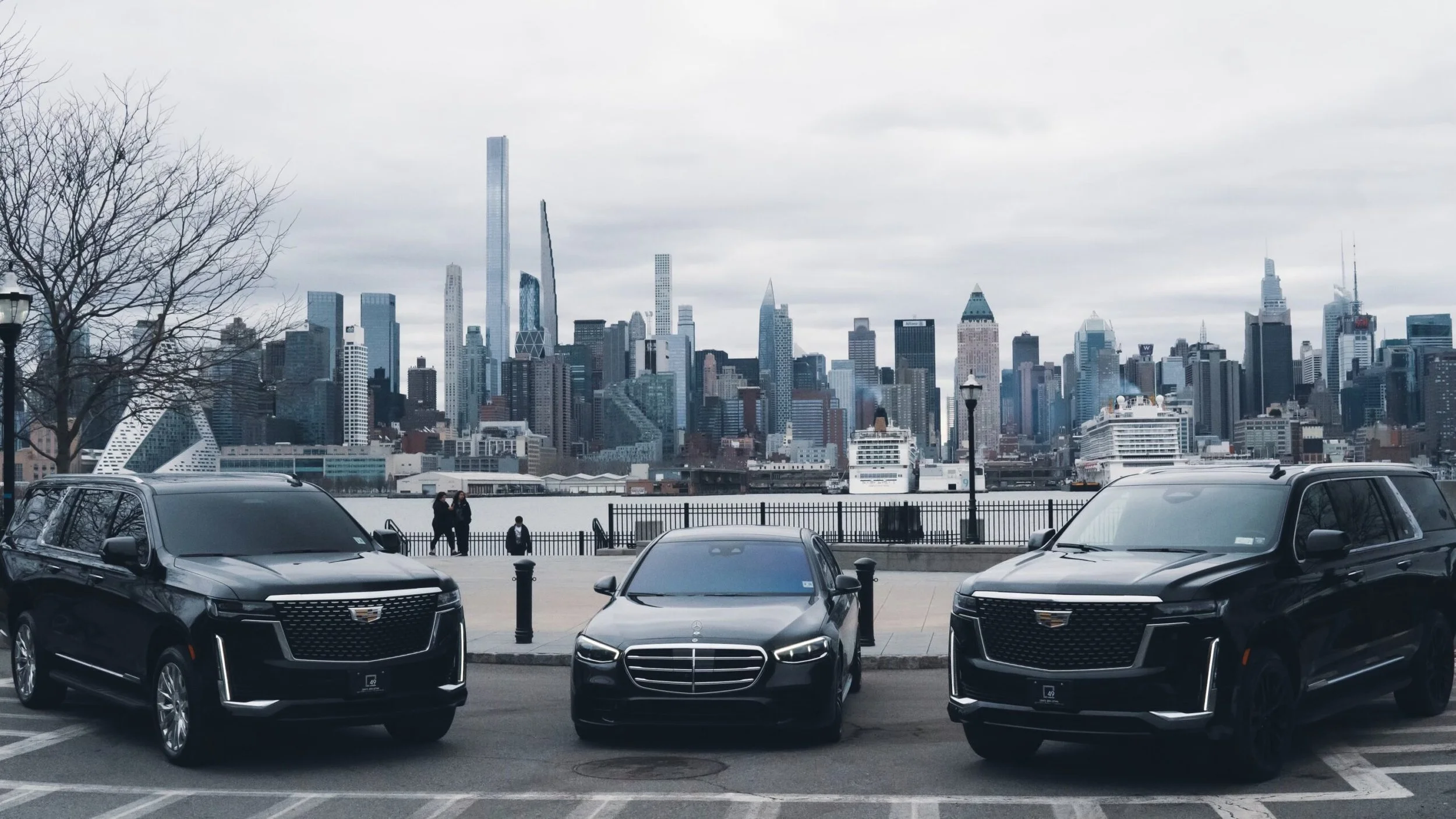 City skyline viewed from a parking lot with several parked cars and two people walking along the sidewalk.