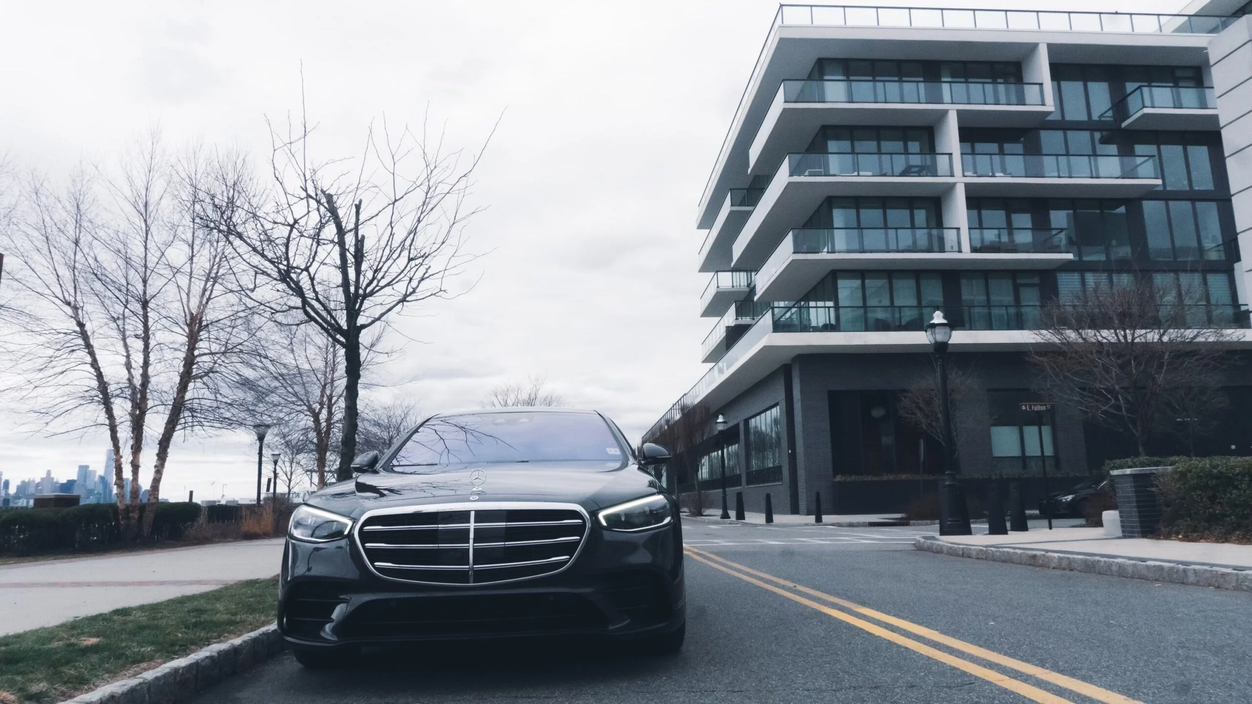 A black Mercedes-Benz car parked on the street near a modern apartment building with glass balconies. Bare trees and a city skyline are in the background under a cloudy sky.