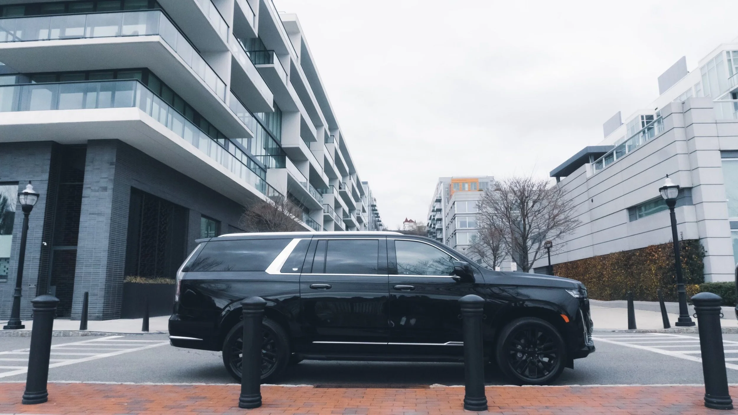 A black SUV parked on the street in front of modern residential buildings with glass balconies and a cloudy sky.