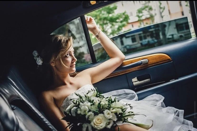 Young woman in bridal dress holding a bouquet, sitting in the back seat of a car, looking out the window.