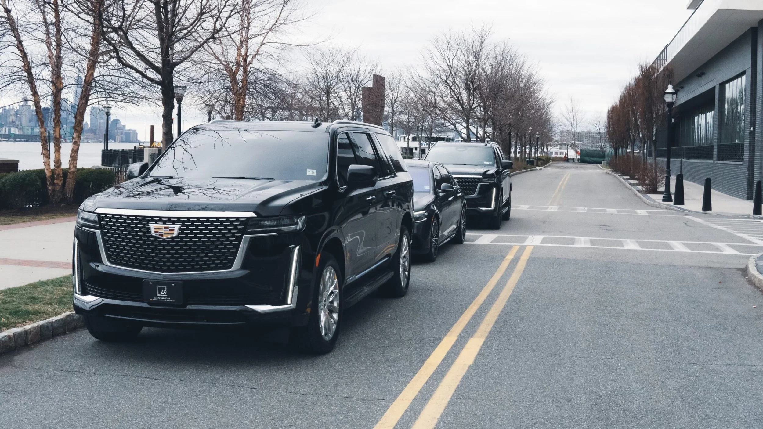 A row of parked black SUVs along a city street near a waterfront, with leafless trees, street lamps, and a city skyline in the background.