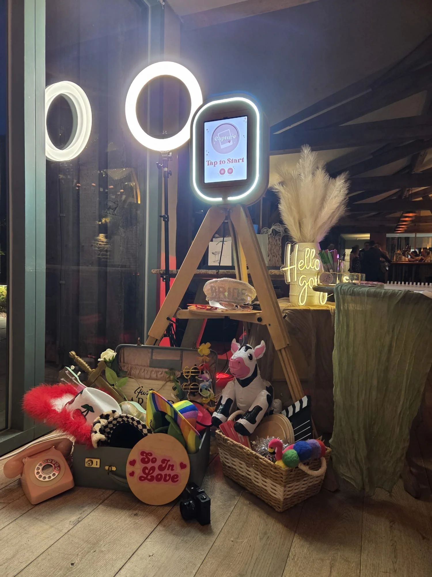 Display of various party accessories and decorations, including a pink toy phone, plush toys, hats, and signs with messages like 'In Love' and 'Hello Galo', set up indoors with a mirror and lights in the background.