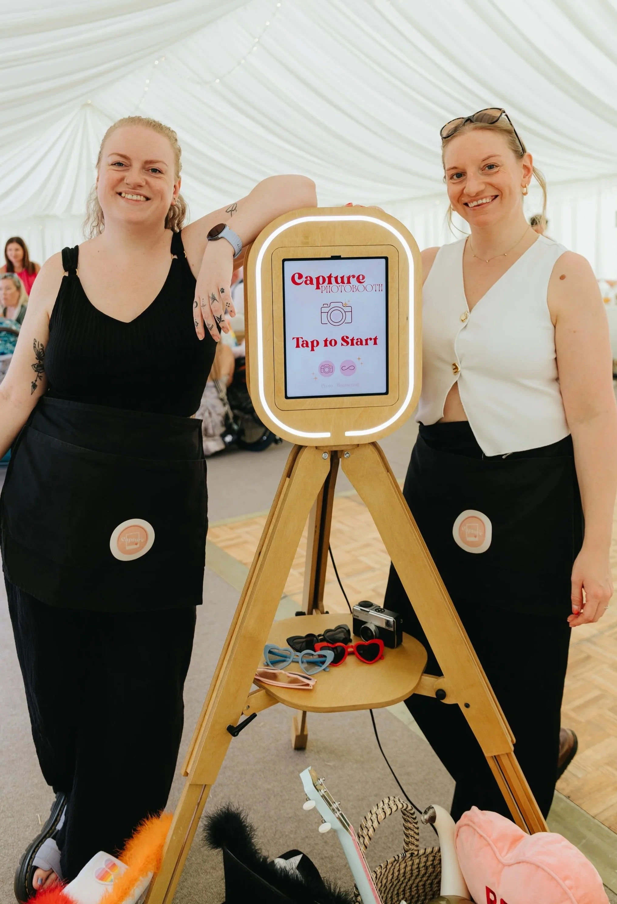 Two smiling women standing next to a wooden photo booth with a sign that says, 'Capture Photo Booth, Tap to Start,' at an indoor event. The women are dressed in black and white tops, and the photo booth has various props like glasses, feathers, and a stuffed animal on a small table.