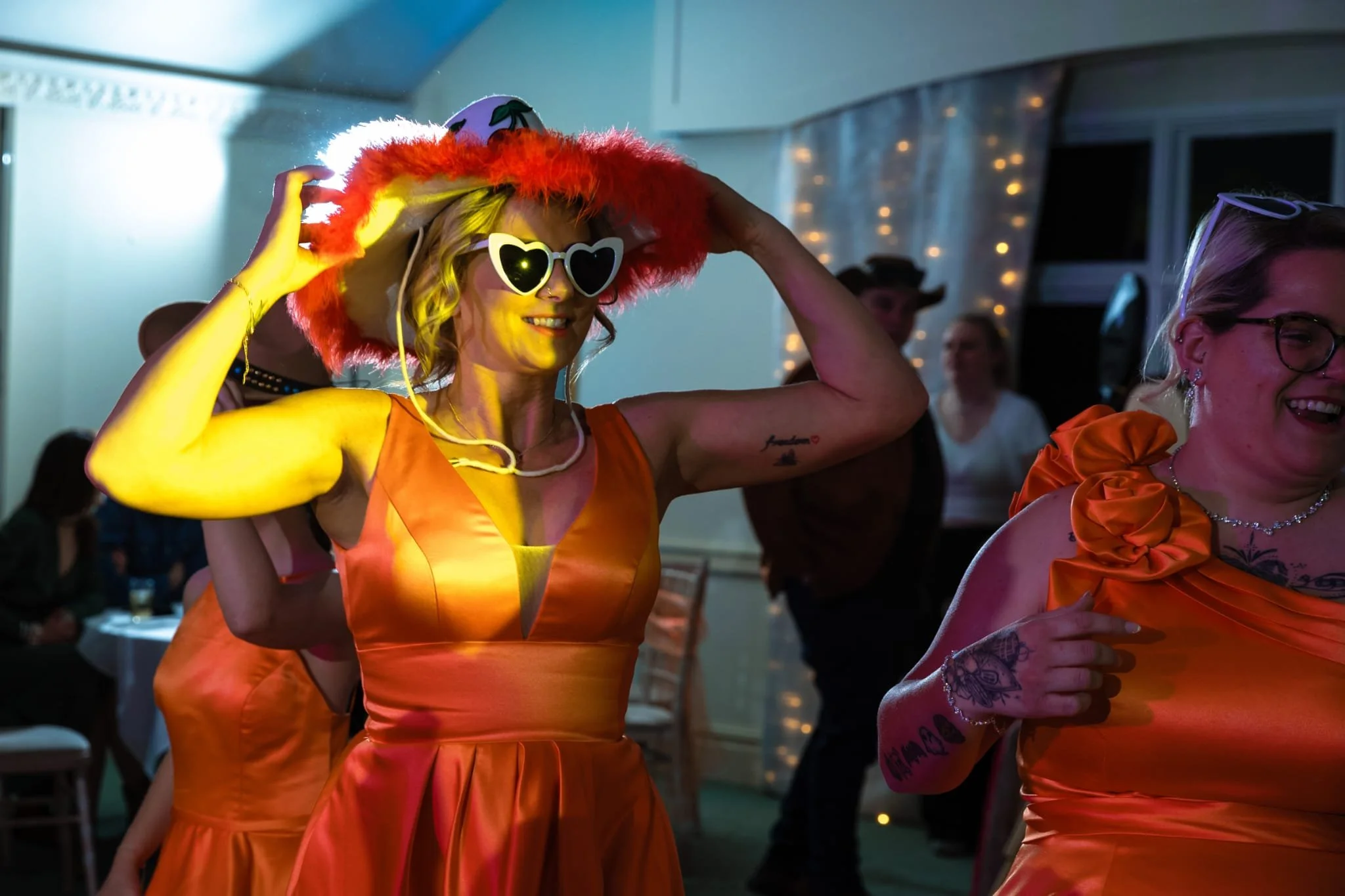 Women dressed in orange at a party, one wearing heart-shaped sunglasses, a red fluffy hat, and a white cake hat, smiling and dancing in a decorated room.